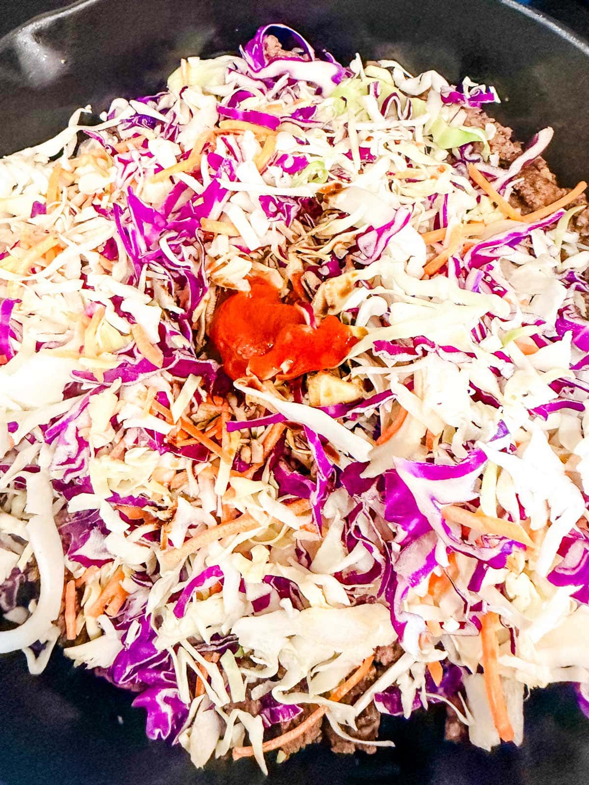 A mix of shredded green and purple cabbage, carrots, and ground meat in a pan, topped with a dollop of red sauce, ready to be cooked.