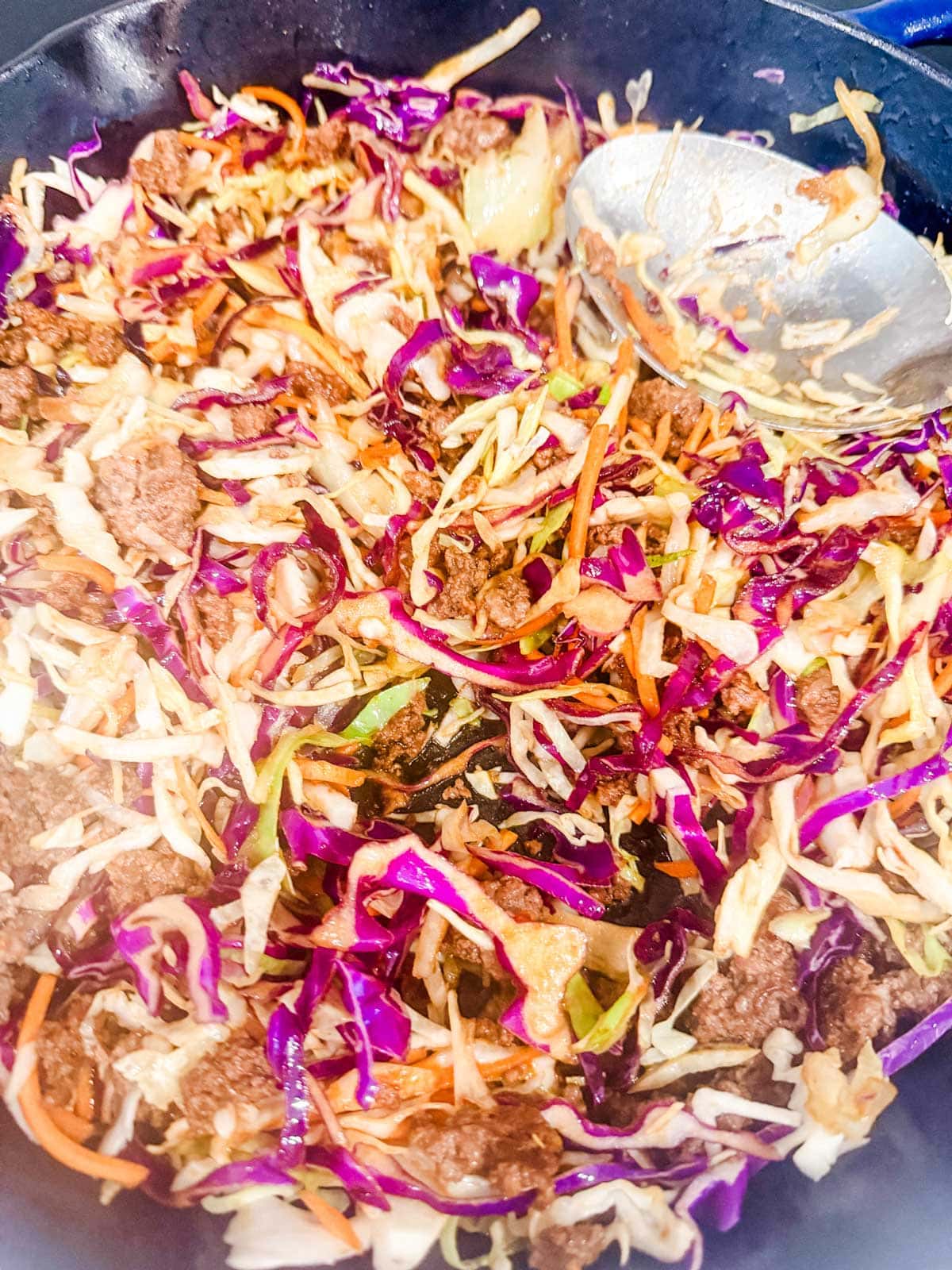 A skillet filled with a colorful mix of cooked ground beef, shredded purple and green cabbage, carrots, and onions, with a metal spoon resting to the side.