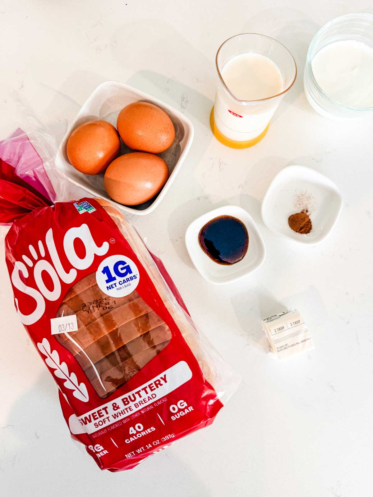 Ingredients for making French toast on a white surface: a loaf of Sola sweet & buttery bread, three eggs, a glass of milk, a bowl of sugar, two sticks of butter, vanilla extract, and a small dish of cinnamon.