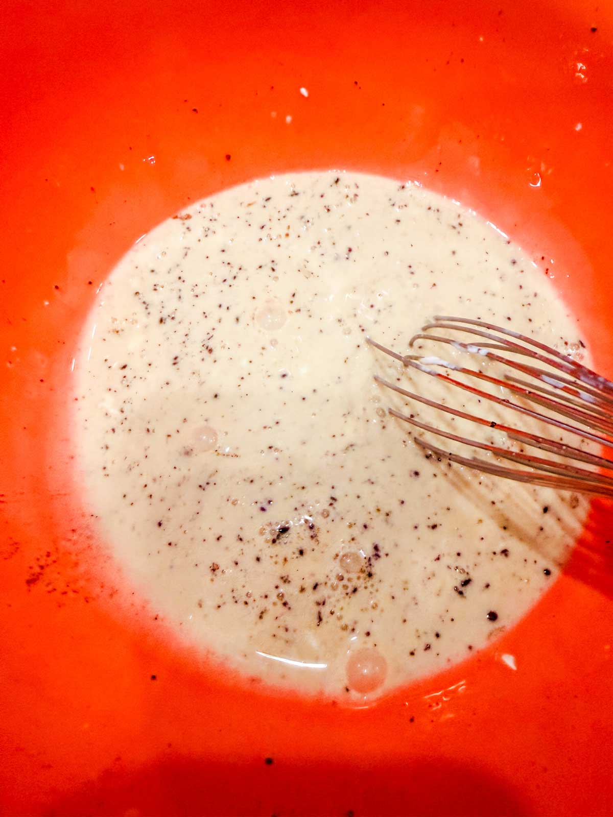 A metal whisk in a red bowl containing a creamy mixture with visible black pepper sprinkled on top.