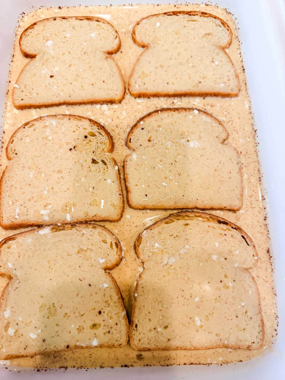 Six slices of bread are soaking in a shallow dish filled with a speckled, creamy liquid, preparing to be made into French toast. The mixture contains visible spices and seasonings.