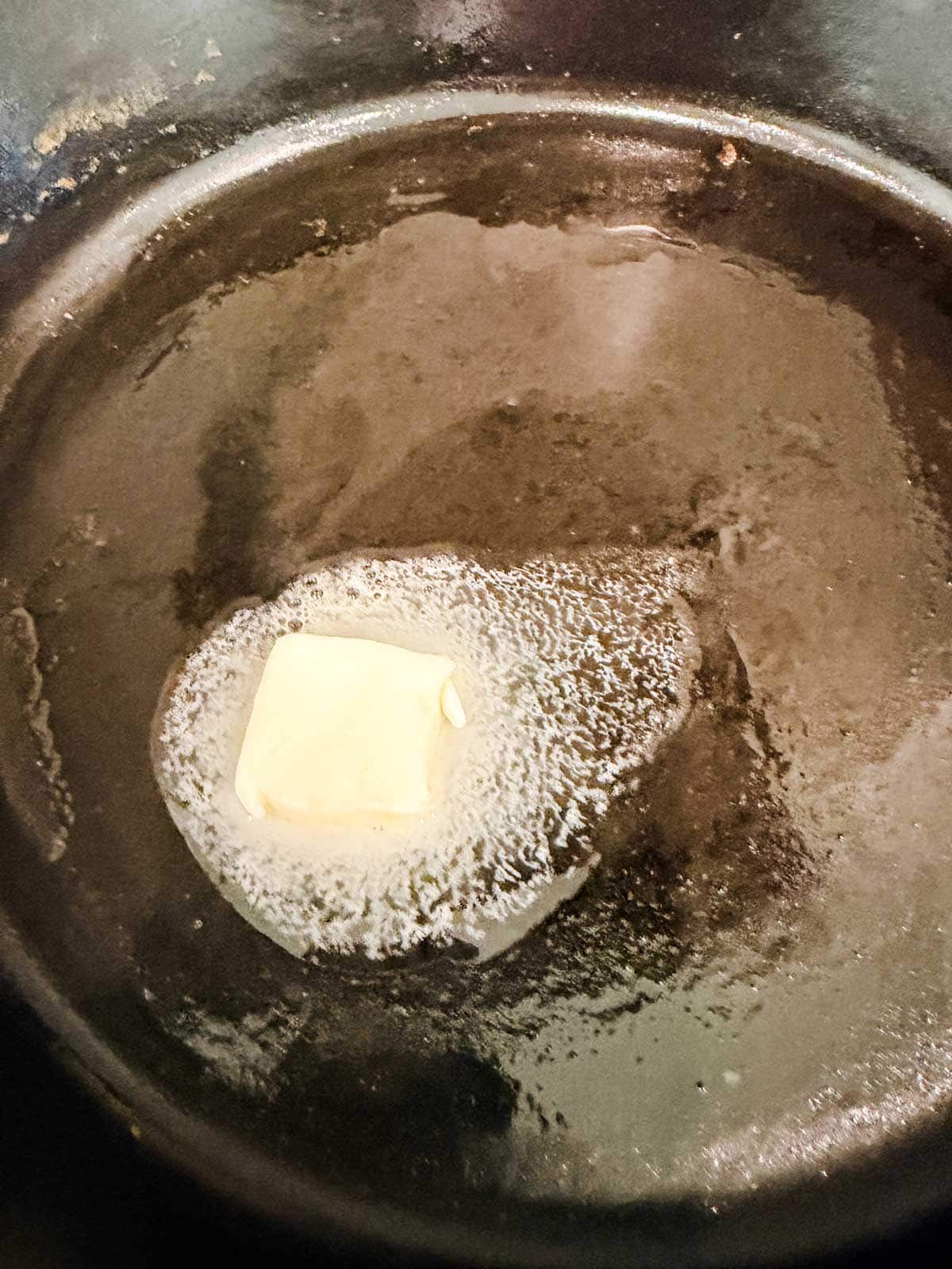 A pat of butter melting in a black skillet, with the butter partially liquefied and starting to spread across the pan.
