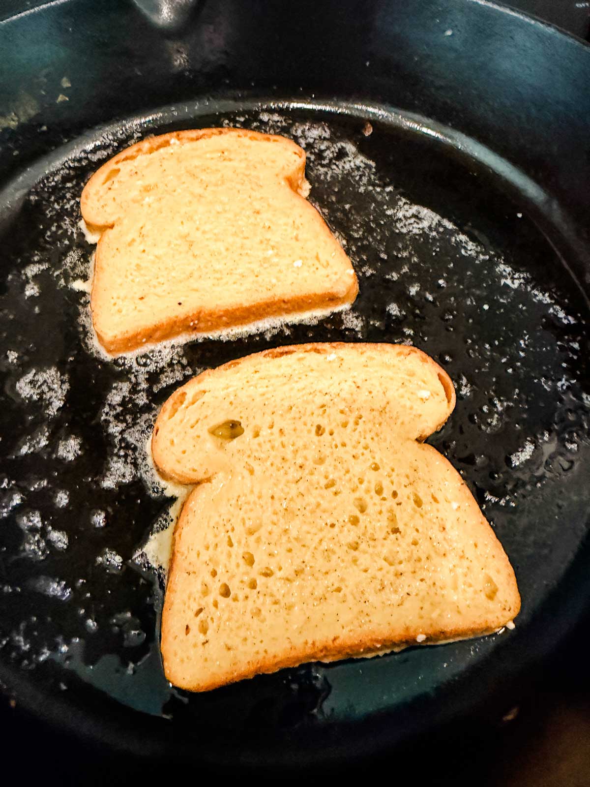 Two slices of bread are being toasted in a black skillet with melted butter, showing a golden-brown color and small bubbles forming around the bread on the pan’s surface.