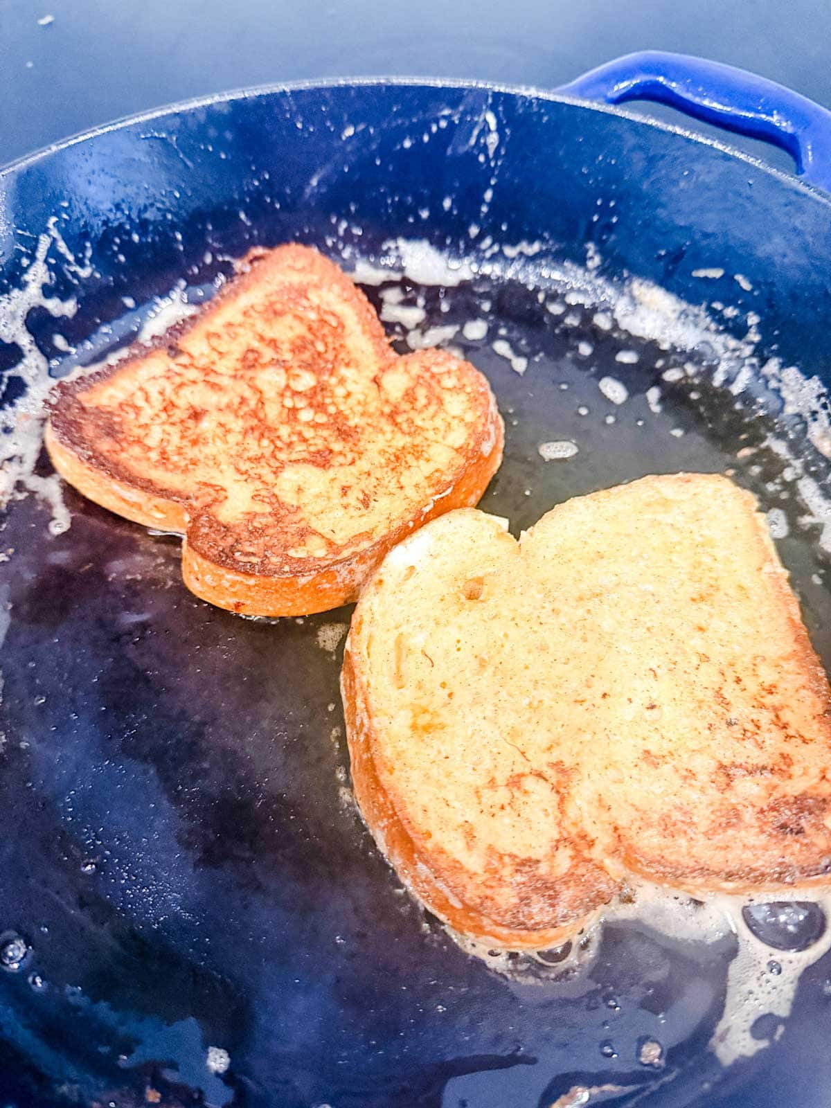 Two slices of French toast are cooking in a blue cast iron skillet, sizzling in melted butter. The bread is golden brown and crispy around the edges.