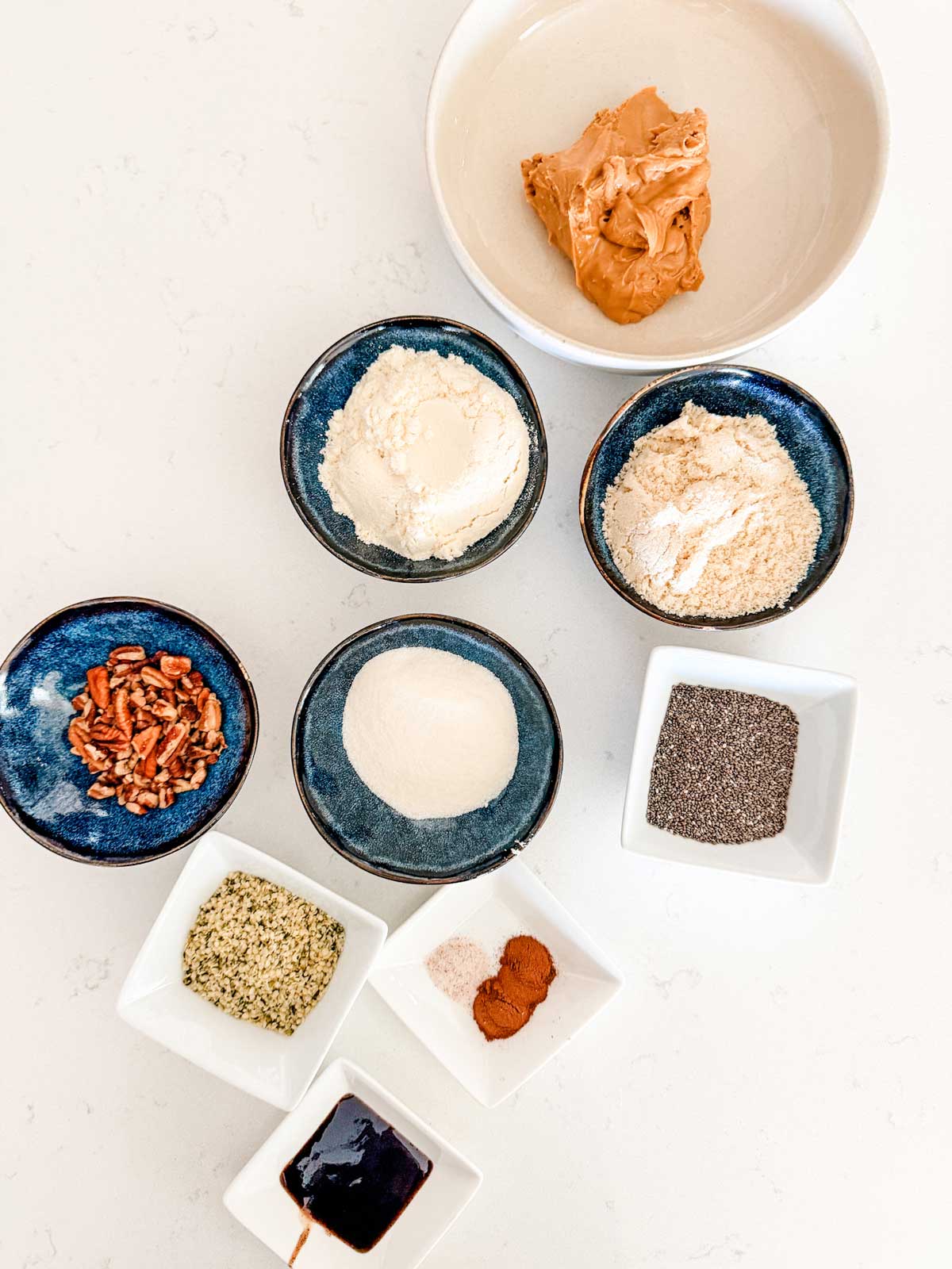Bowls and dishes containing peanut butter, keto sweetener, almond flour, chopped pecans, ground flaxseed, chia seeds, hemp seeds, vanilla, salt, and cinnamon are arranged on a white surface.