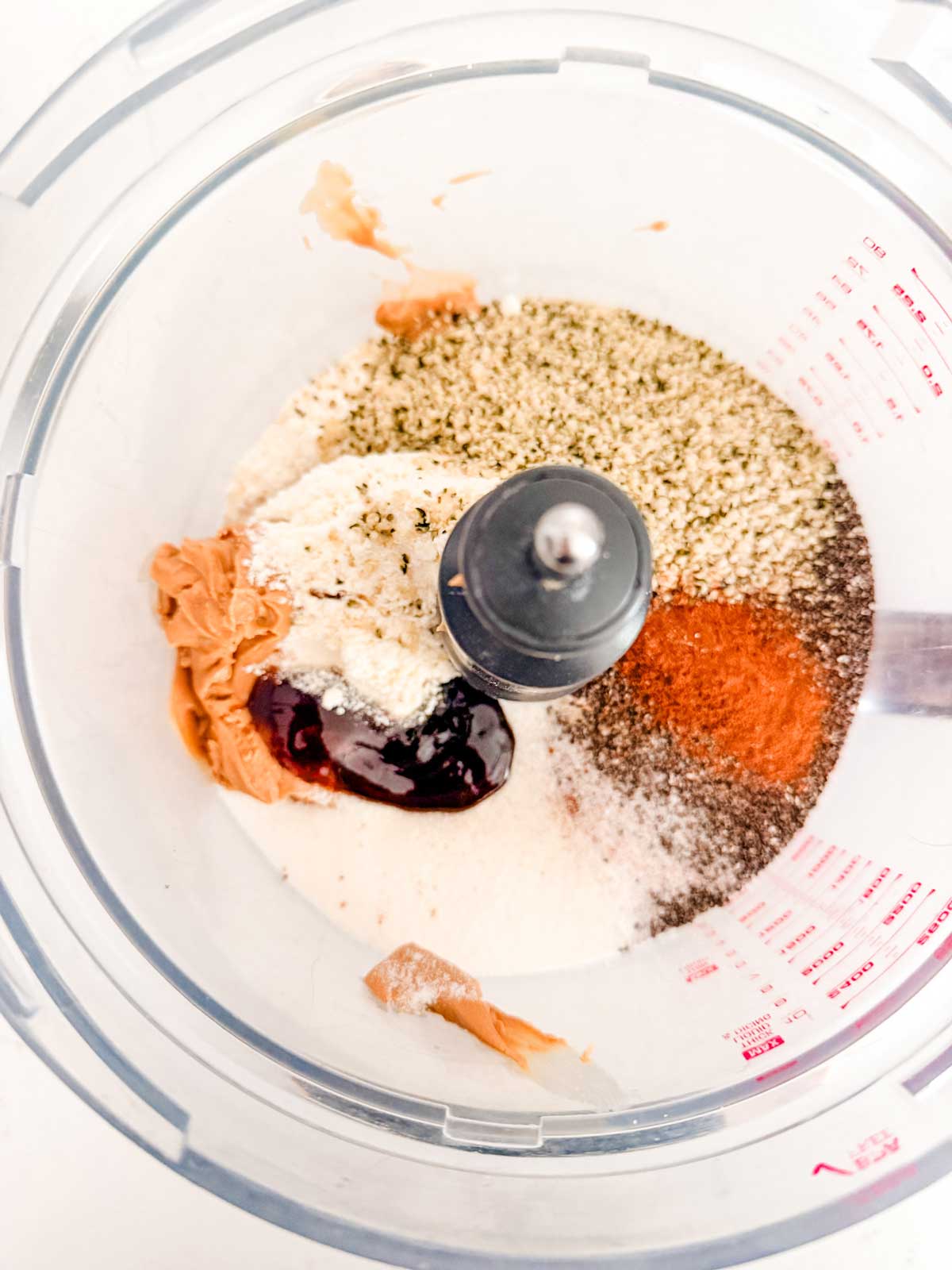 A food processor bowl filled with various ingredients, including peanut butter, vanilla, ground spices, seeds, and powders, ready to be mixed together.