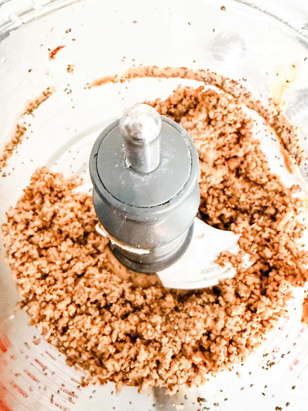 A close-up of ground nuts, protein powder, seeds, sweetener and cinnamon in a food processor bowl, showing a coarse, crumbly mixture around the metal blade.