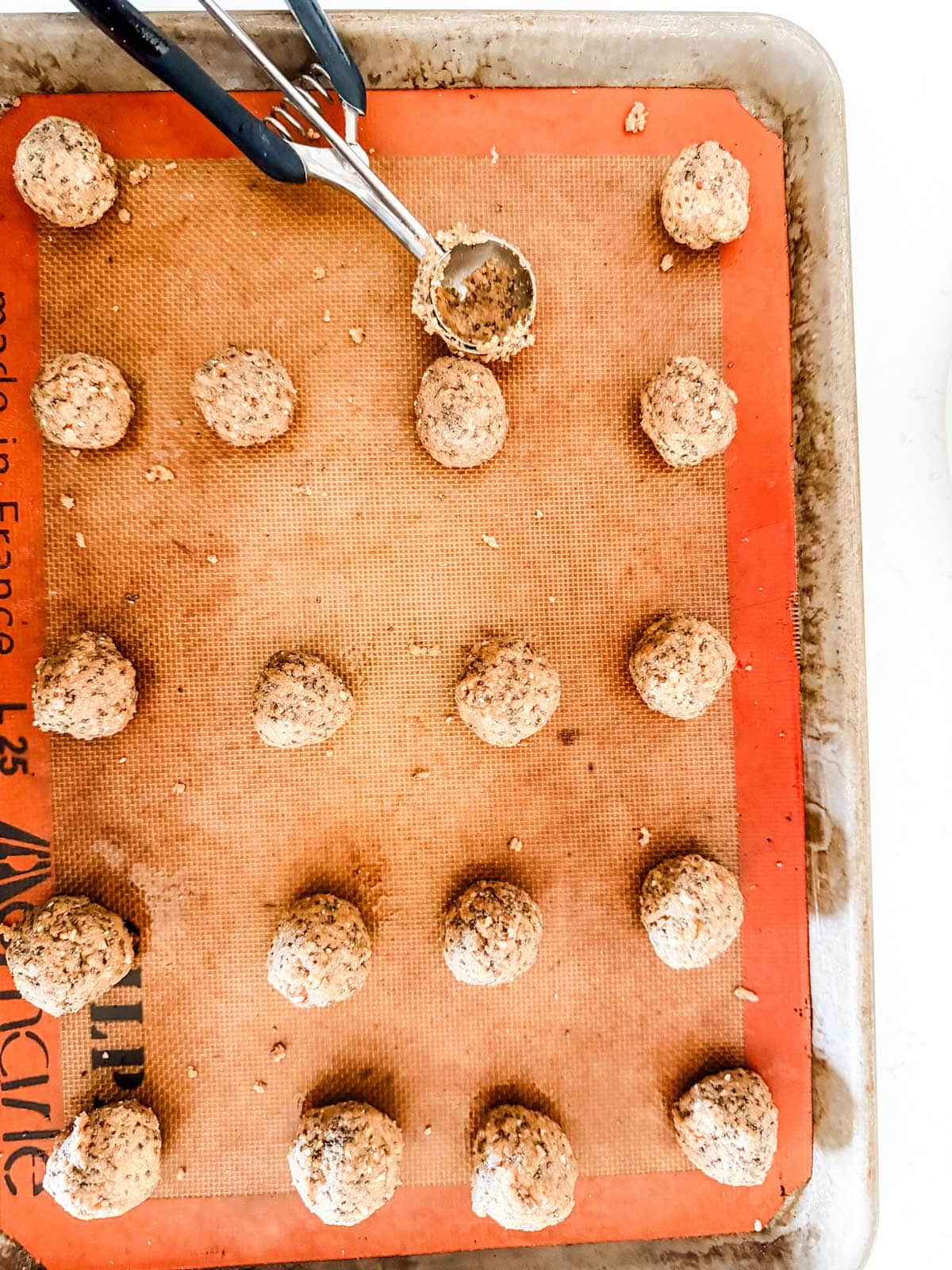 A baking sheet lined with a silicone mat holds evenly spaced unbaked balls. A metal cookie scoop with dough inside rests at the top of the tray.