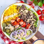 A keto Big Mac bowl filled with shredded cheddar cheese, cooked ground beef, sliced pickles, cherry tomatoes, red onion, lettuce, and creamy dressing, topped with black sesame seeds. Cherry tomatoes and fresh parsley sit nearby.