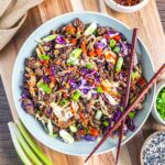 A keto egg roll in a bowl with stir-fried ground meat, shredded cabbage, green onions, and sriracha sauce, garnished with sesame seeds. Served with chopsticks on a wooden board, plus small bowls of green onions and sesame seeds on the side.