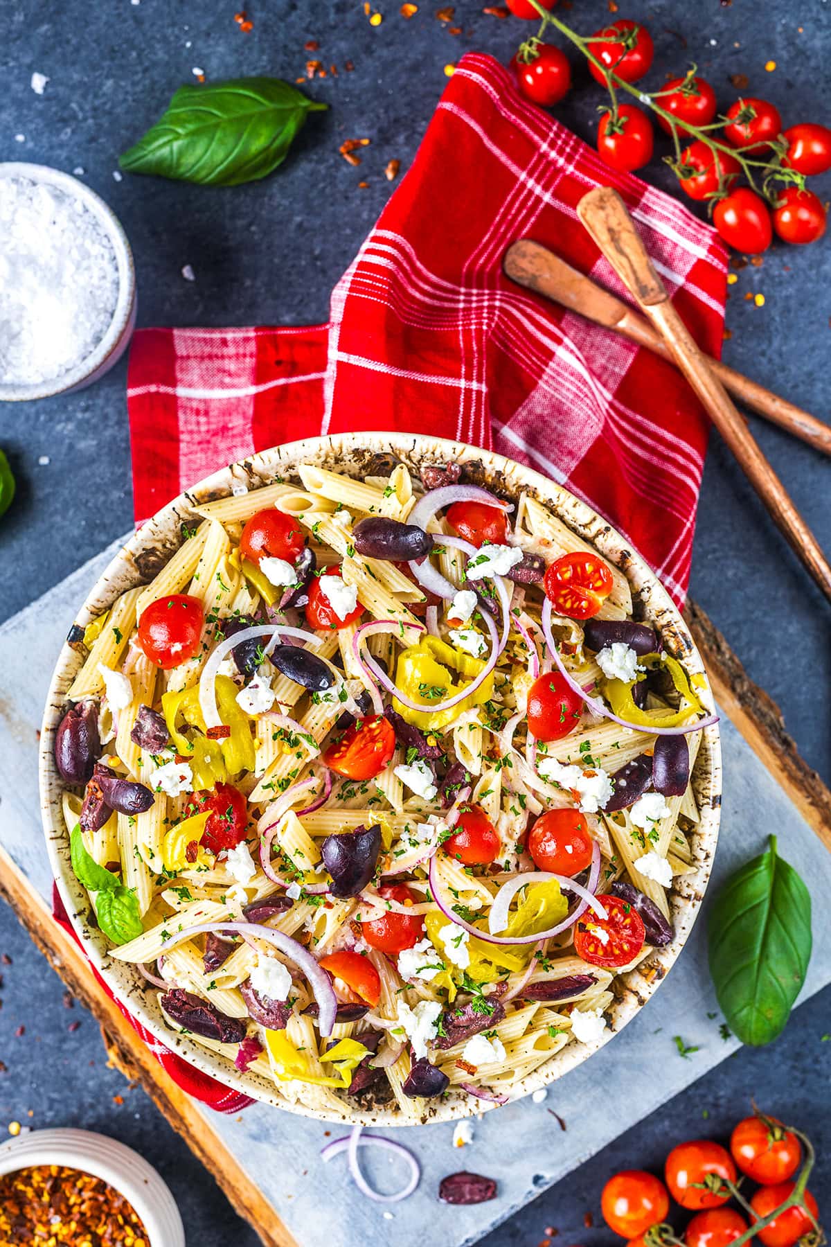 A bowl of pasta salad with cherry tomatoes, black olives, red onion, pepperoncini, and feta cheese sits on a red plaid cloth, surrounded by fresh basil, tomatoes, and wooden utensils on a gray surface.