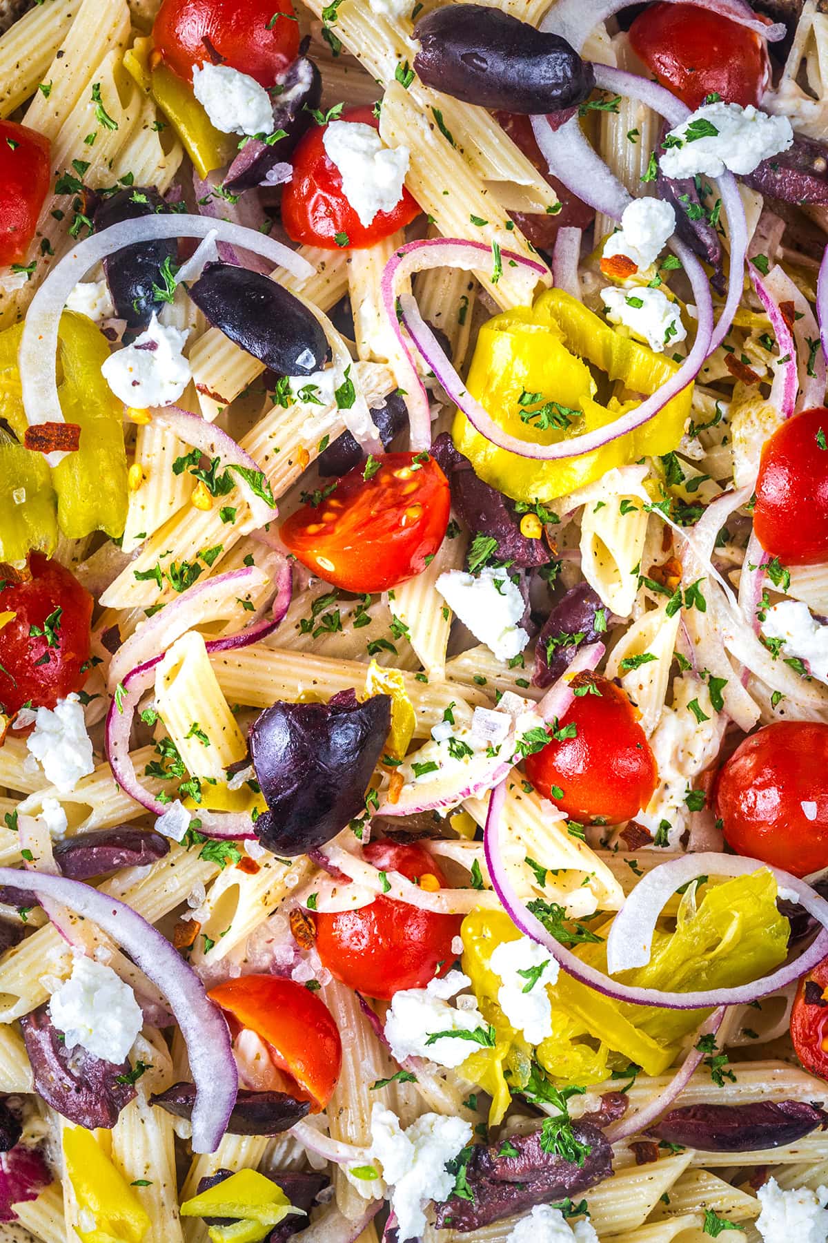 Colorful close-up of penne pasta salad with cherry tomatoes, black olives, red onion slices, pepperoncini, crumbled feta cheese, and chopped parsley, mixed together for a vibrant Mediterranean dish.