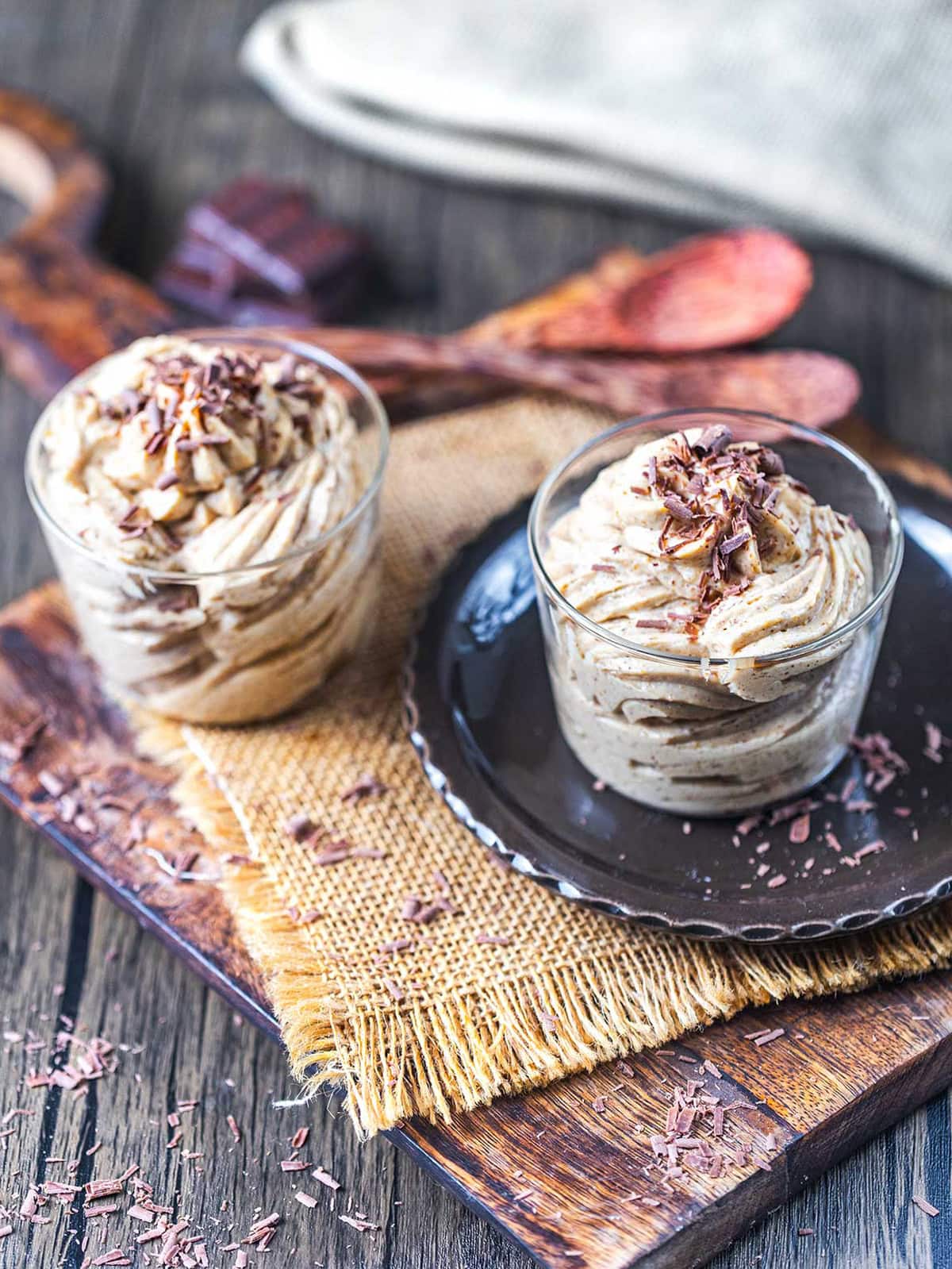 Two glass cups filled with creamy dessert topped with chocolate shavings sit on a dark plate and rustic wooden board, with a burlap cloth and wooden utensils in the background.