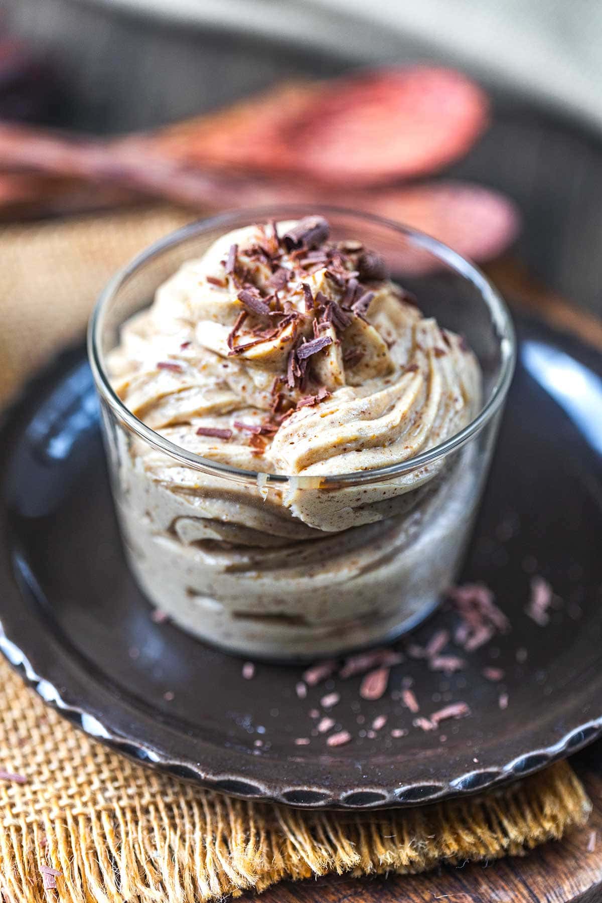 A glass cup filled with creamy, swirled mousse topped with chocolate shavings sits on a dark plate with a woven mat underneath. Wooden spoons are blurred in the background.