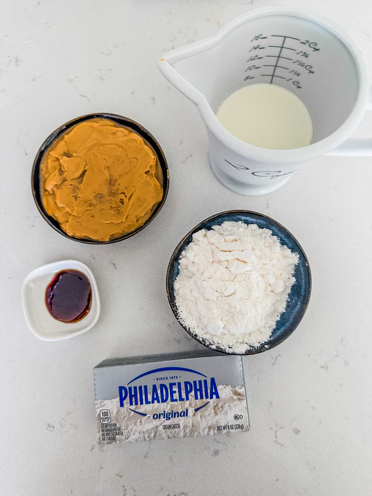 Four ingredients on a white surface: a bowl of peanut butter, a measuring cup with milk, a bowl of flour, a small dish with vanilla extract, and a block of Philadelphia cream cheese.