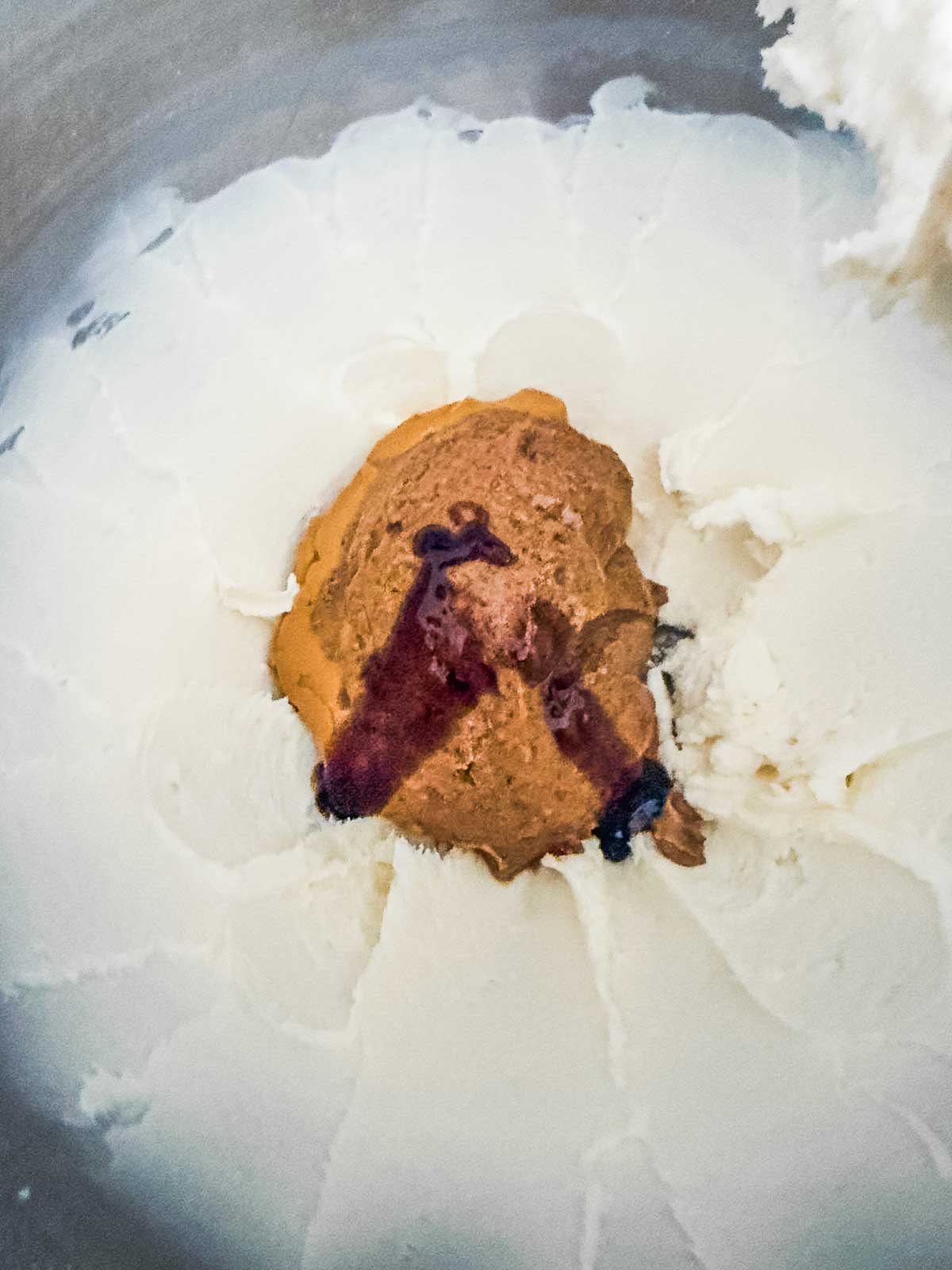 A close-up of a mixing bowl containing cream cheese, a dollop of peanut butter, and a drizzle of vanilla extract, ready to be mixed together.