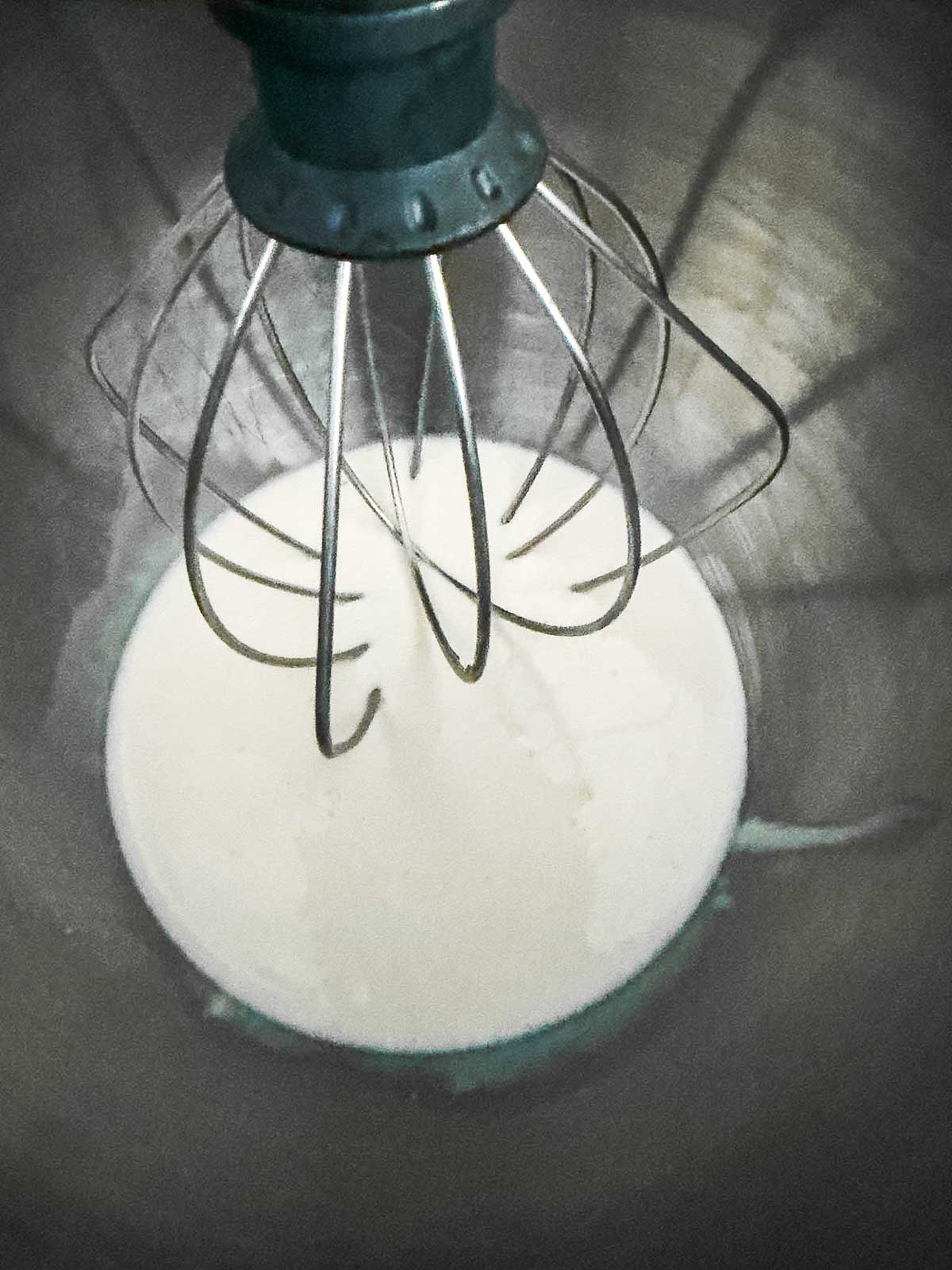 A close-up view of a metal whisk attachment in a stand mixer, whipping cream in a mixing bowl.