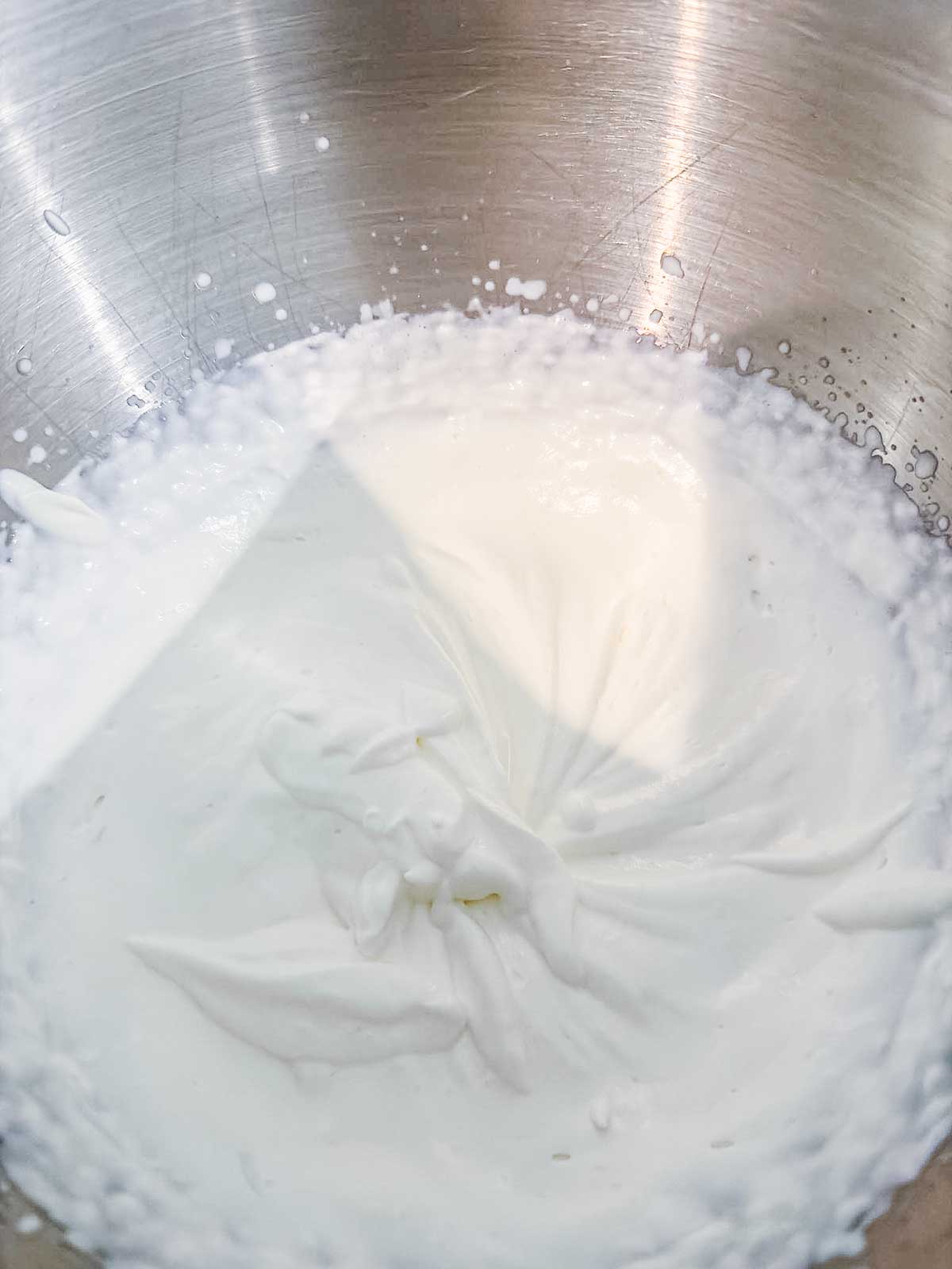 A close-up of whipped cream with soft peaks forming in a stainless steel mixing bowl.