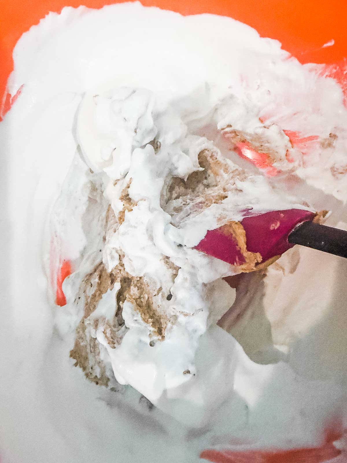 A close-up of a mixing bowl with white whipped cream and a brown peanut butter mixture being folded together using a purple spatula. The bowl is orange.