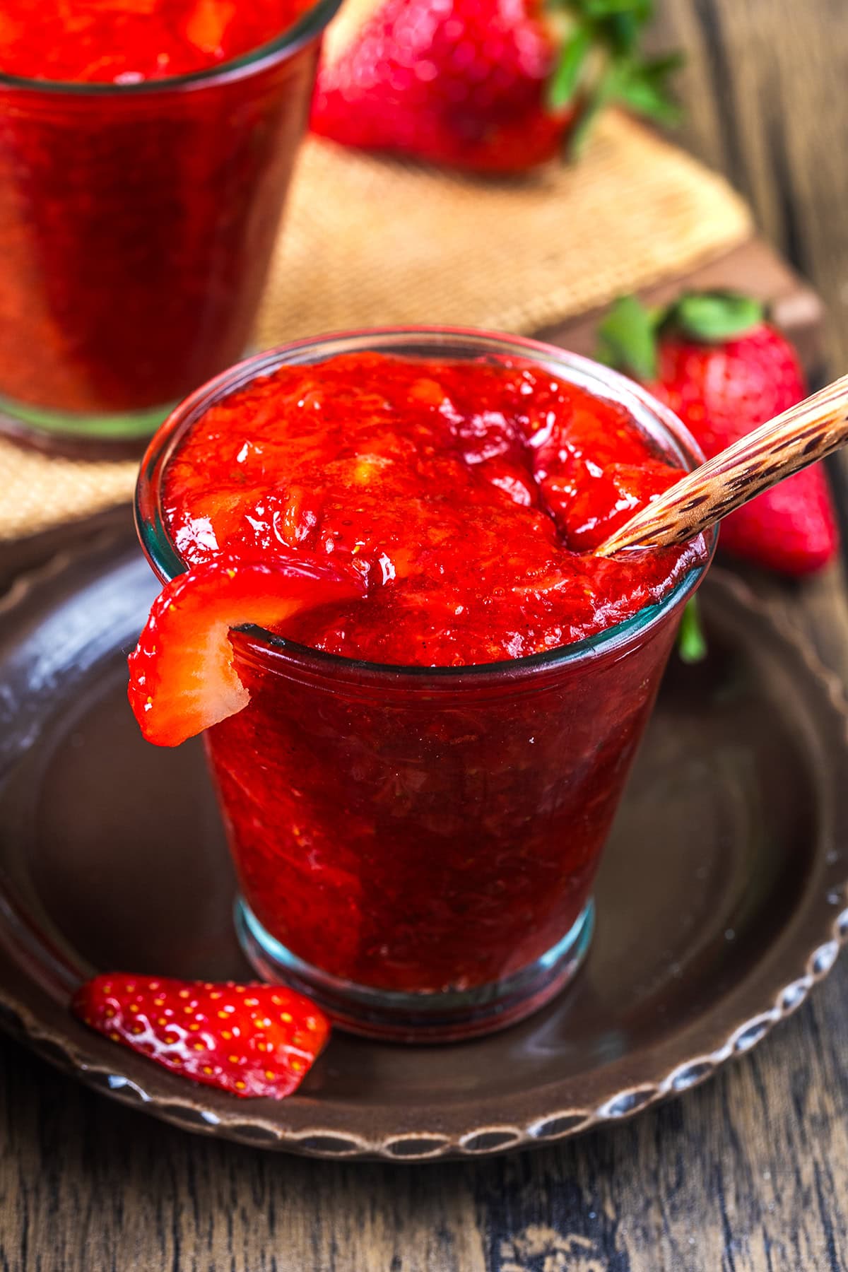 A glass filled with thick, homemade strawberry jam, garnished with a fresh strawberry slice on the rim, sits on a dark plate with a wooden spoon and more strawberries in the background.