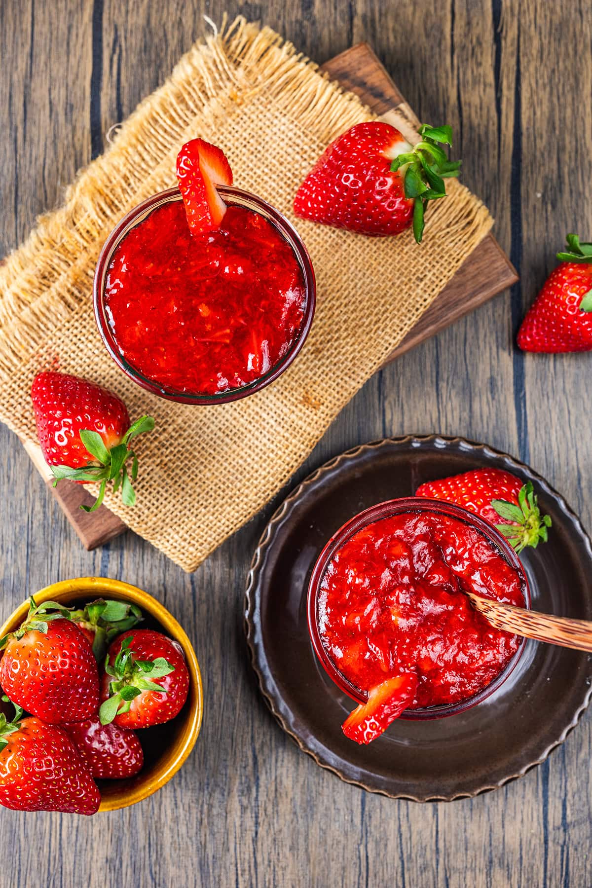 Two glass jars filled with strawberry jam, garnished with fresh strawberries, sit on a rustic wooden surface. Extra strawberries are scattered around, with one small bowl of whole strawberries nearby.