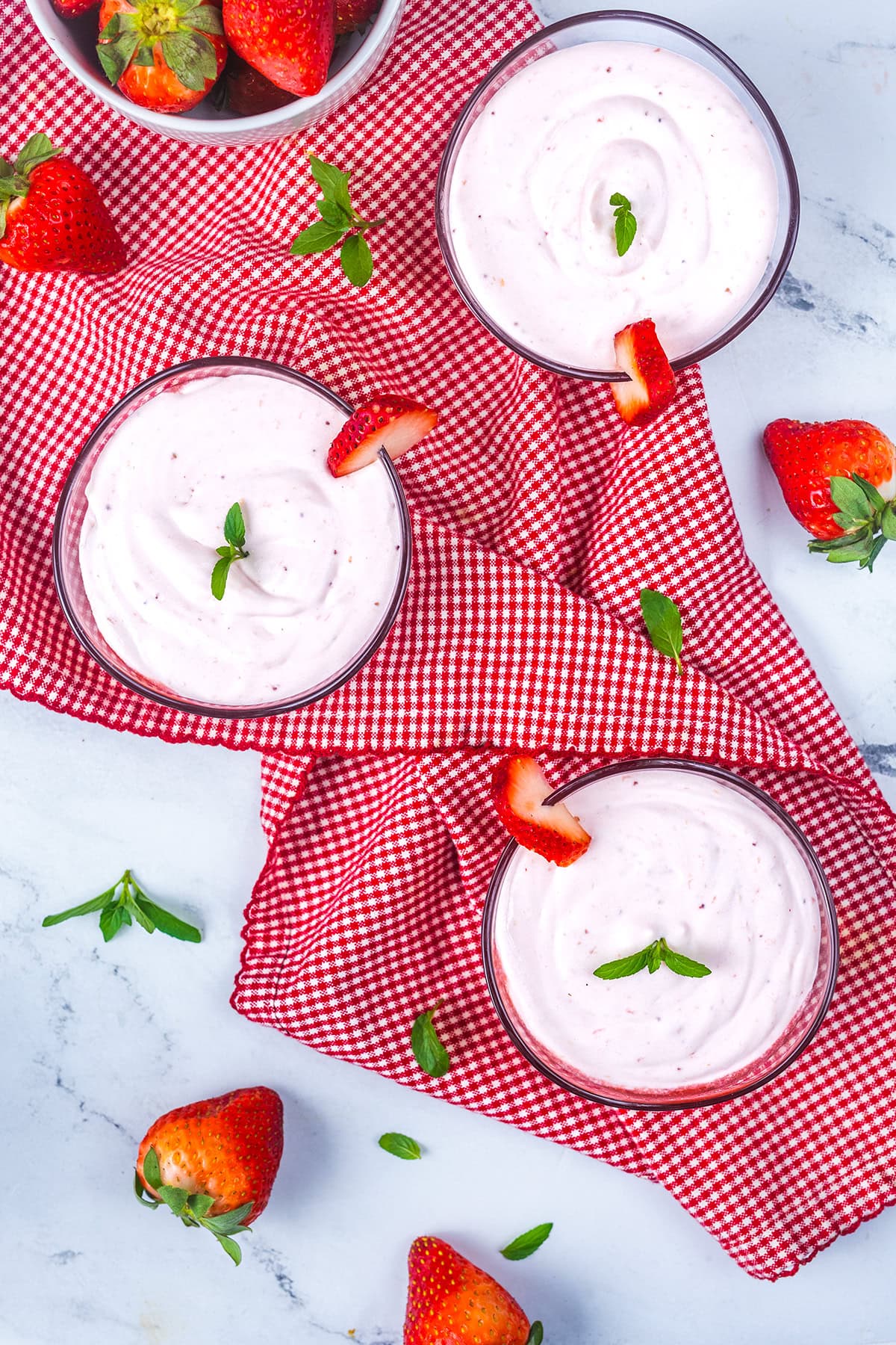 Three glass bowls of strawberry mousse with mint leaves and strawberry slices on top are placed on a red checkered cloth, surrounded by fresh strawberries on a white marble surface.