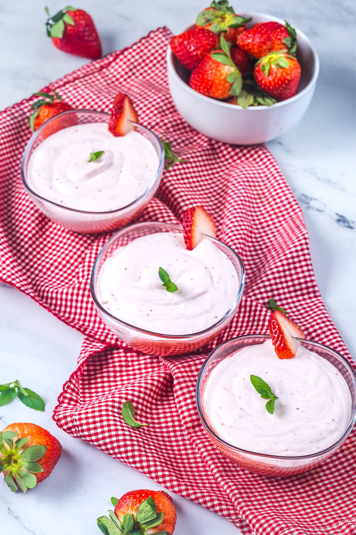 Three glass bowls of strawberry mousse garnished with fresh strawberry slices and mint leaves sit on a red checkered cloth, with a bowl of whole strawberries in the background.