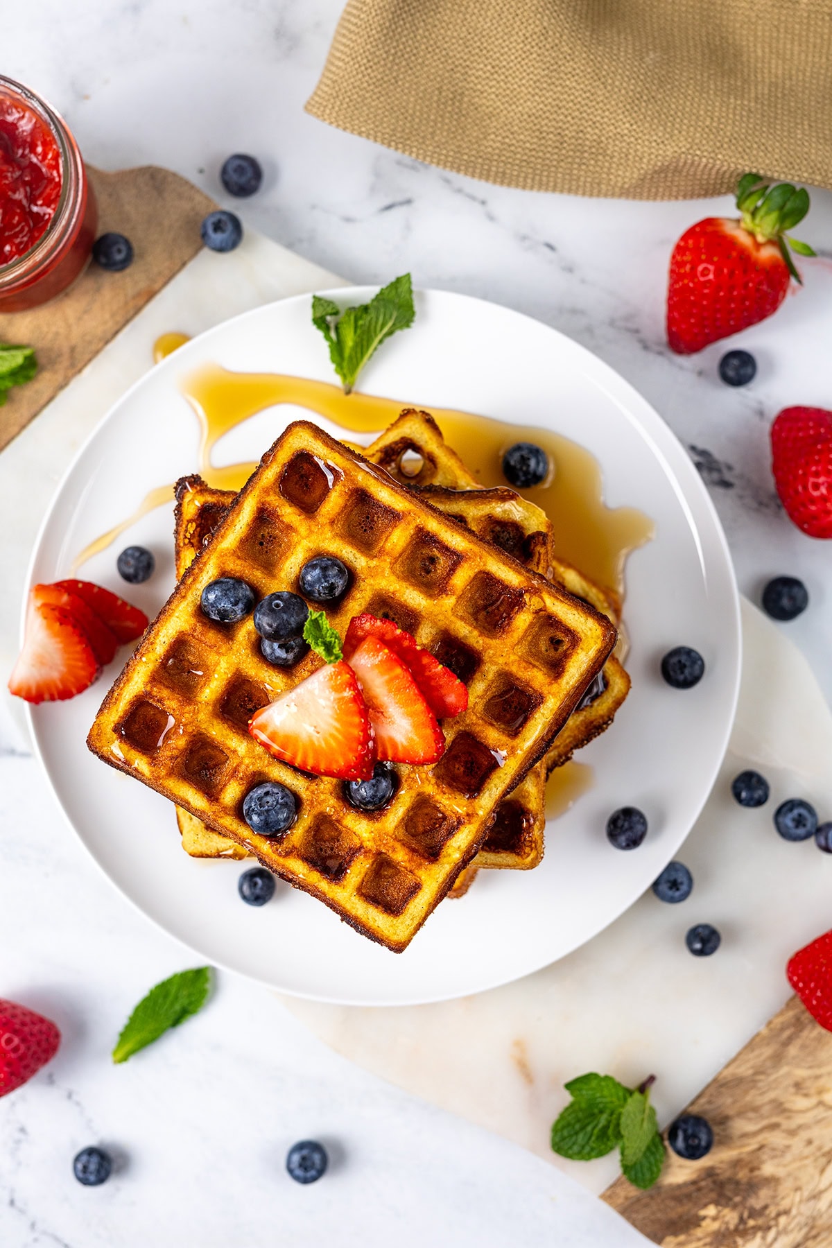 A plate of waffles topped with sliced strawberries, blueberries, and syrup, surrounded by scattered berries and mint leaves on a white marble surface. A jar of jam, a strawberry, and a napkin are nearby.