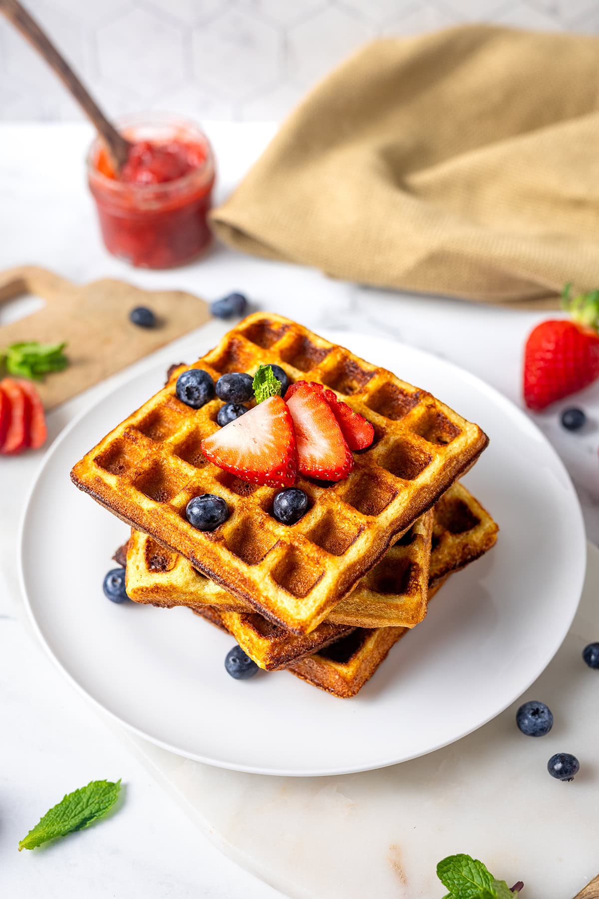 Two golden waffles stacked on a white plate, topped with sliced strawberries, blueberries, and a mint leaf. A jar of jam, a wooden board with strawberries, and a beige napkin are in the background.