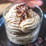 A clear glass filled with creamy, swirled mousse topped with chocolate shavings, sitting on a dark plate.