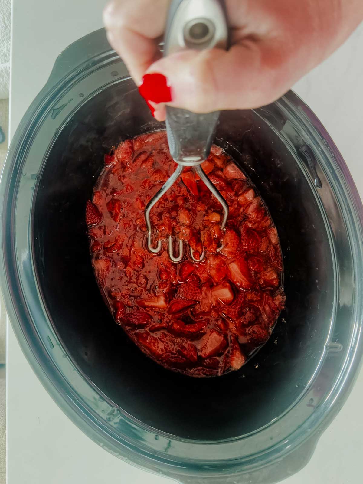 A hand with red nail polish uses a potato masher to mash cooked strawberries in a black slow cooker.