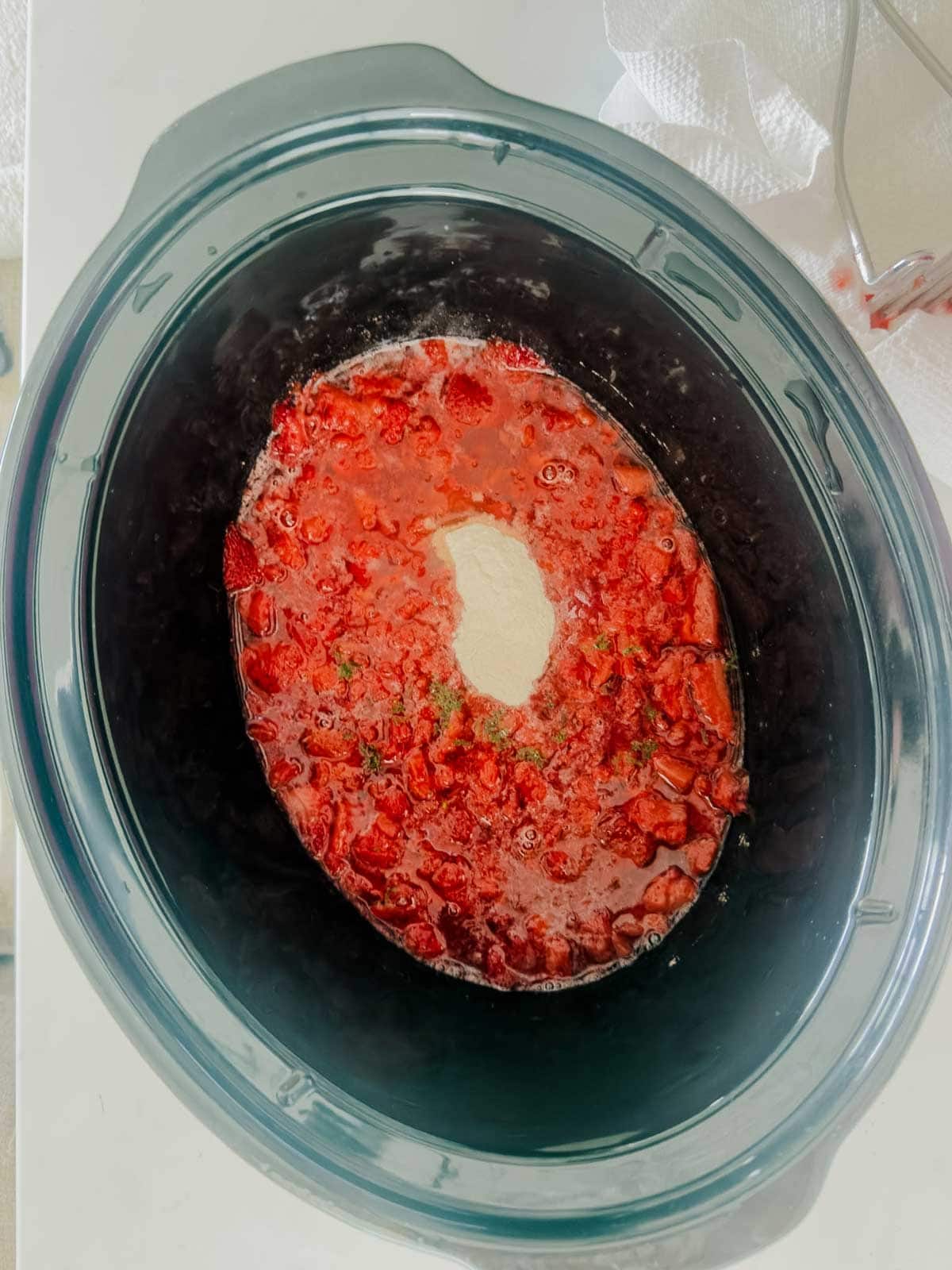 Overhead view of a slow cooker filled with chopped strawberries, sugar, and other ingredients forming a mixture with a mound of white powder in the center, ready for cooking. A paper towel and fork are visible nearby.