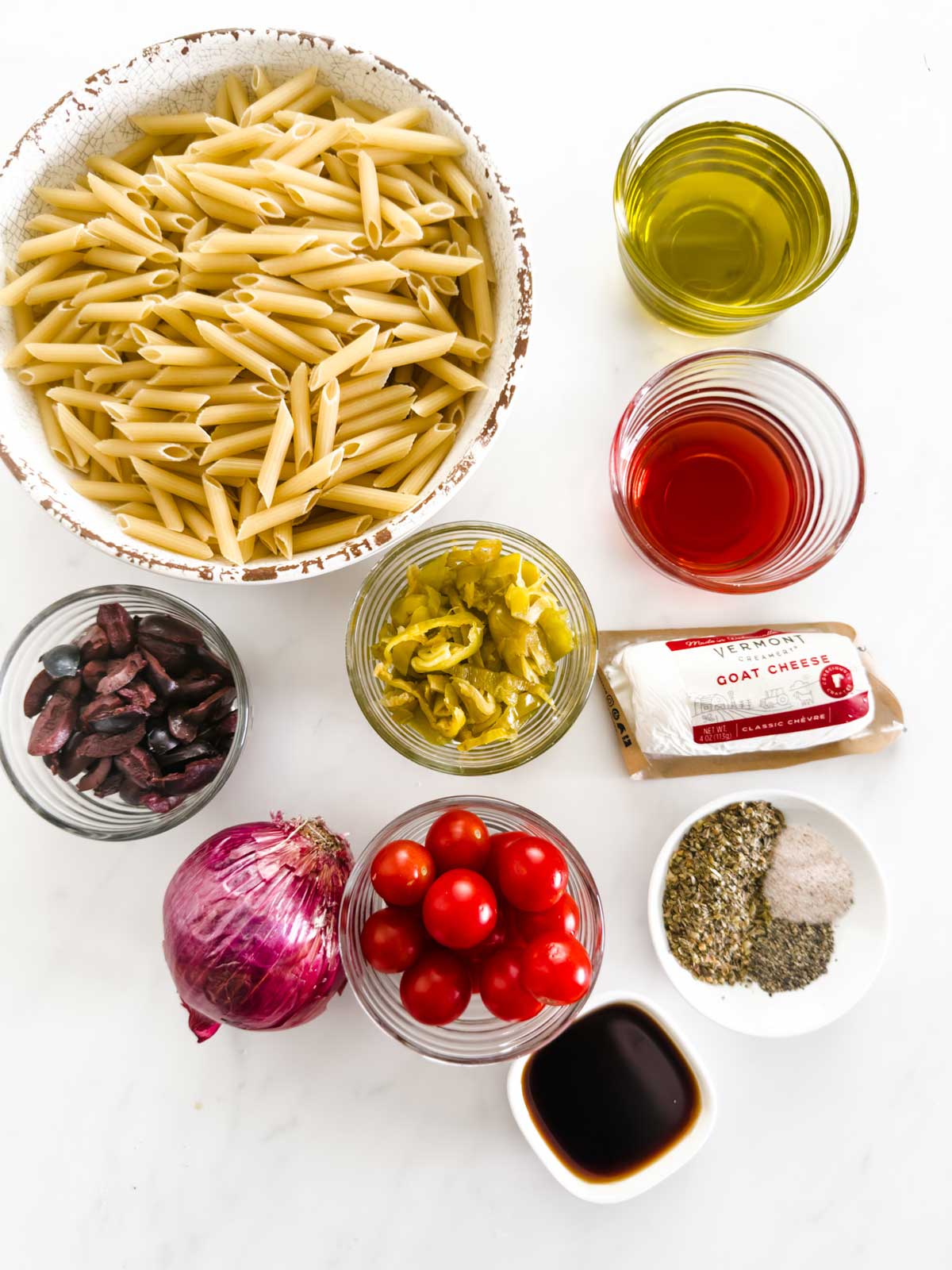 Overhead view of ingredients for a pasta dish: uncooked penne, olive oil, red wine vinegar, kalamata olives, pepperoncini, goat cheese, red onion, cherry tomatoes, dried herbs, salt, pepper, and balsamic vinegar.