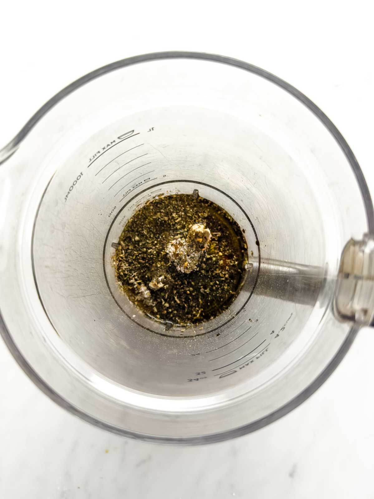 A glass measuring cup with oil, black pepper, and seasonings at the bottom, viewed from above, with a metal whisk inside.