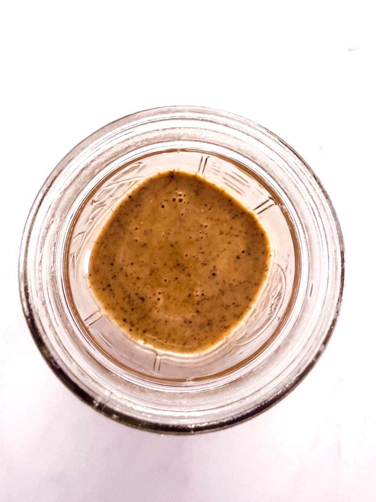 A top-down view of a glass jar containing a creamy, brown sauce or dressing with visible black specks, set against a white background.