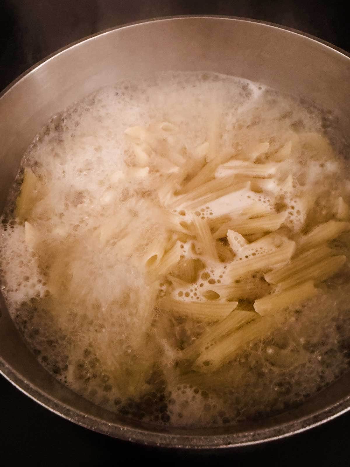 A pot of penne pasta boiling in water with bubbles and steam rising, as the pasta cooks on a stovetop.