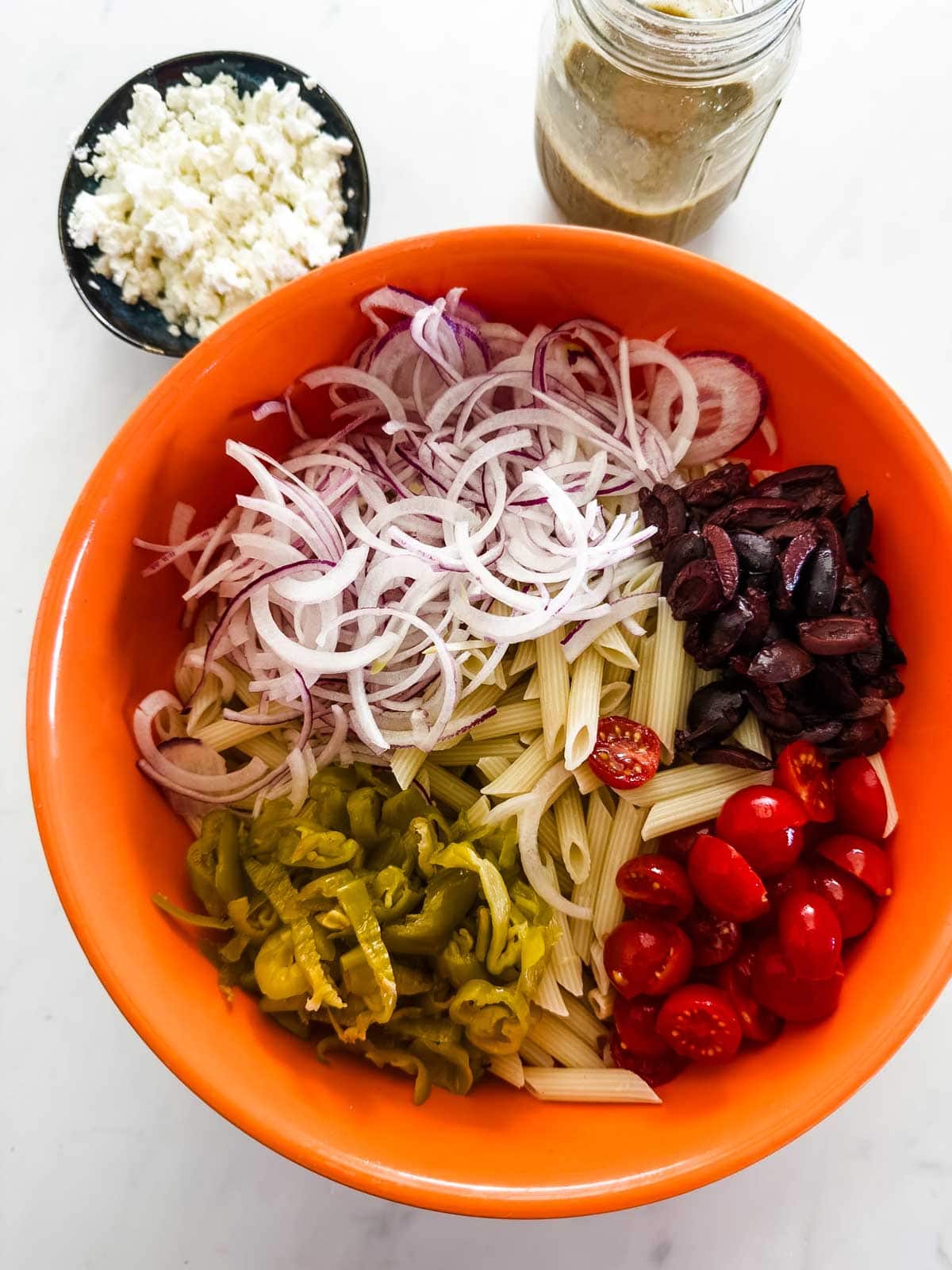 An orange bowl with penne pasta, sliced red onions, Kalamata olives, grape tomatoes, and pickled peppers. Nearby are a small bowl of crumbled cheese and a jar of dressing on a white surface.