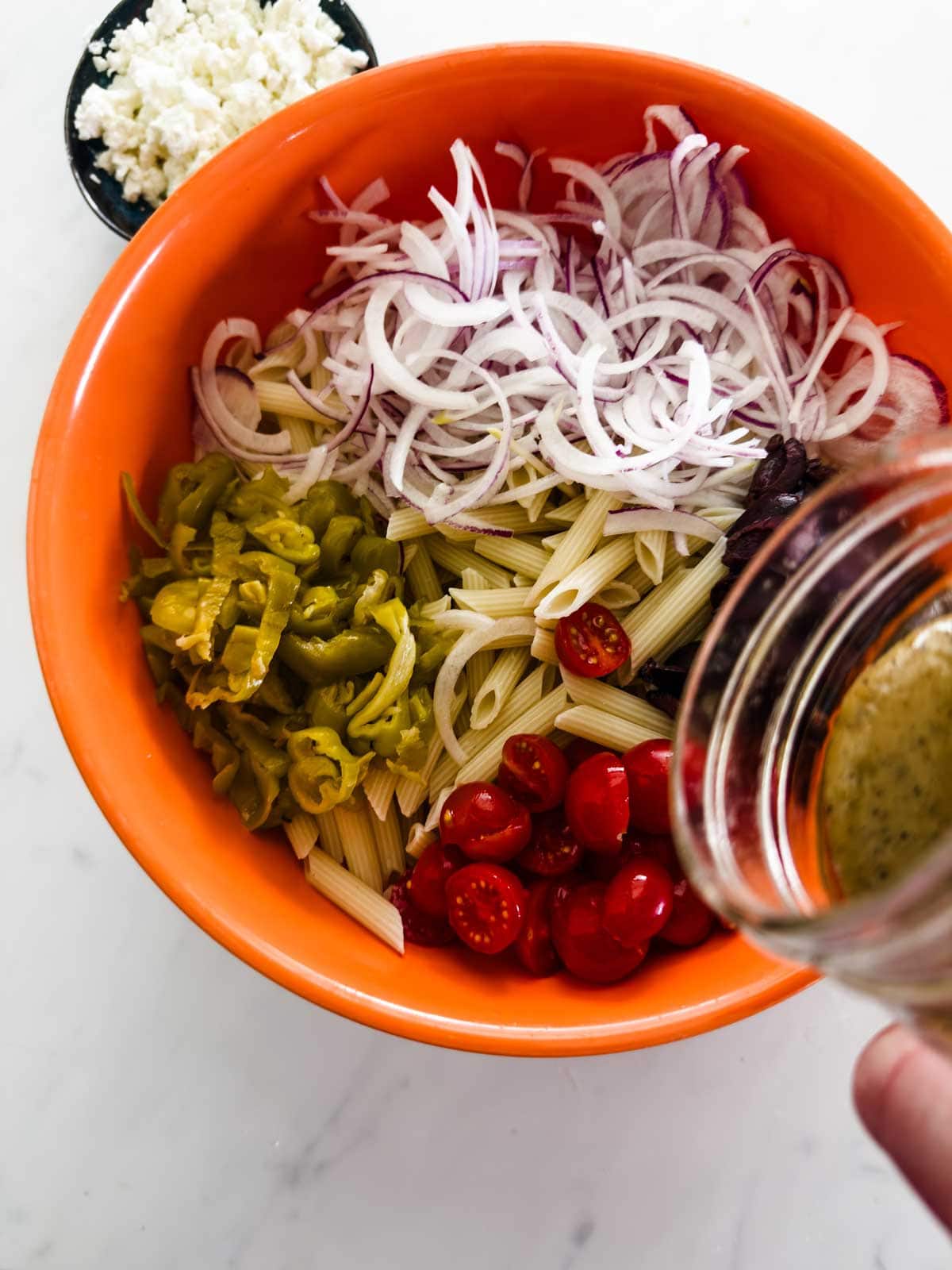 An orange bowl filled with penne pasta, sliced red onions, cherry tomatoes, pepperoncini, and olives. A hand is pouring dressing over the ingredients. A small dish of crumbled cheese is nearby on the white surface.