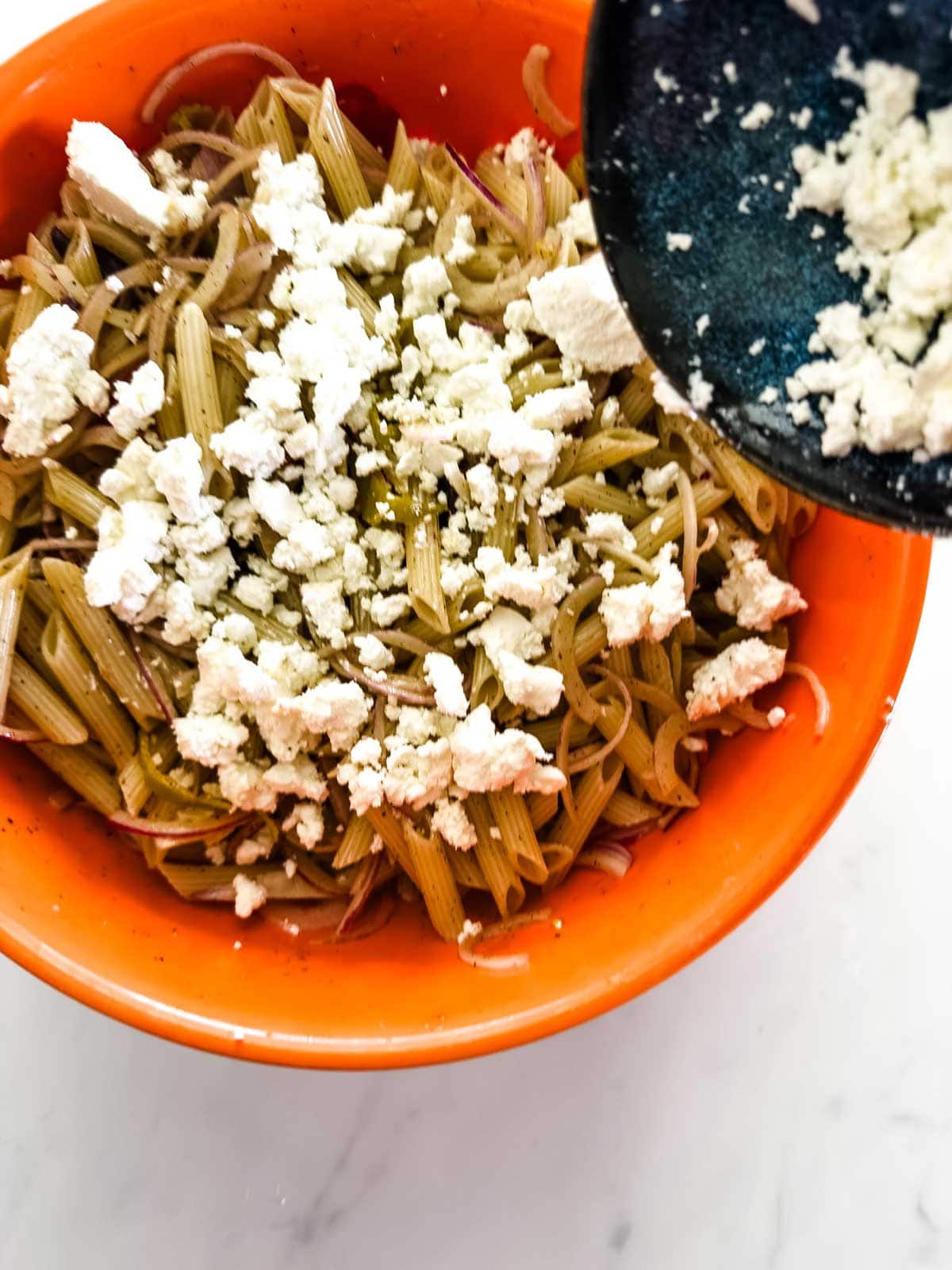 A bright orange bowl filled with cooked penne pasta and thinly sliced red onions, topped with crumbled white cheese being added from a dark blue plate.