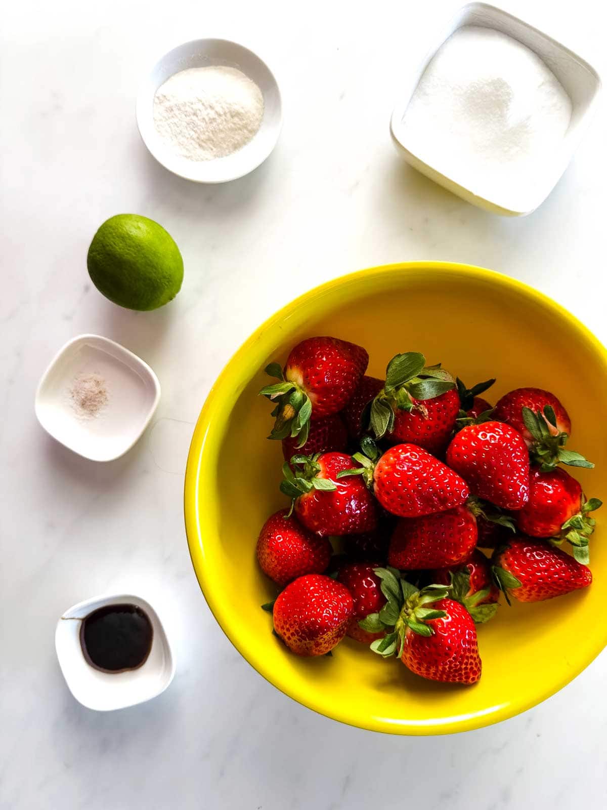 A yellow bowl filled with fresh strawberries sits on a white surface, surrounded by small bowls containing keto sweetener, pectin, vanilla extract, and salt, with a whole lime nearby.