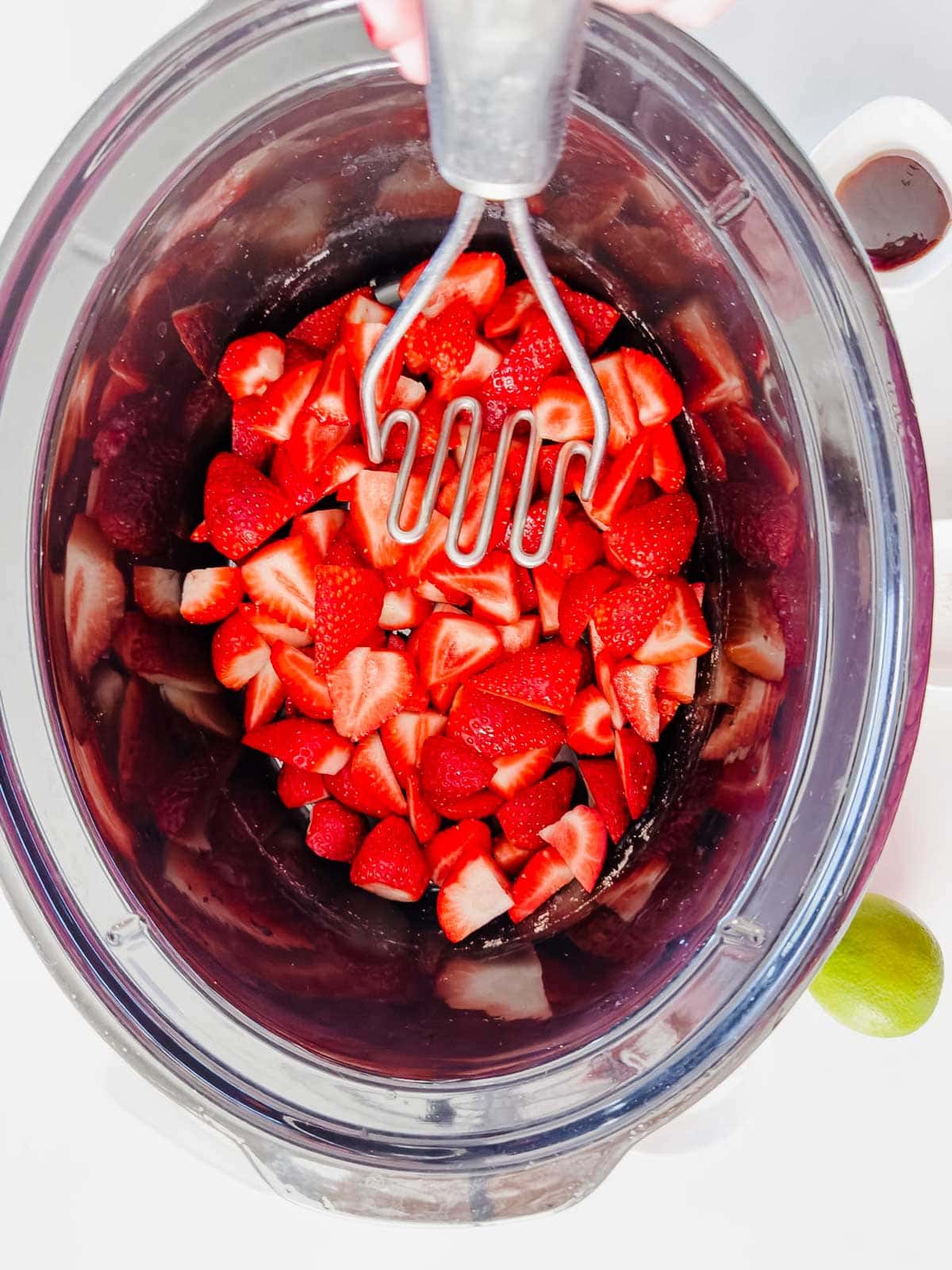 A top-down view of a bowl filled with chopped strawberries being mashed with a metal potato masher; a lime and a small bowl of liquid are nearby.