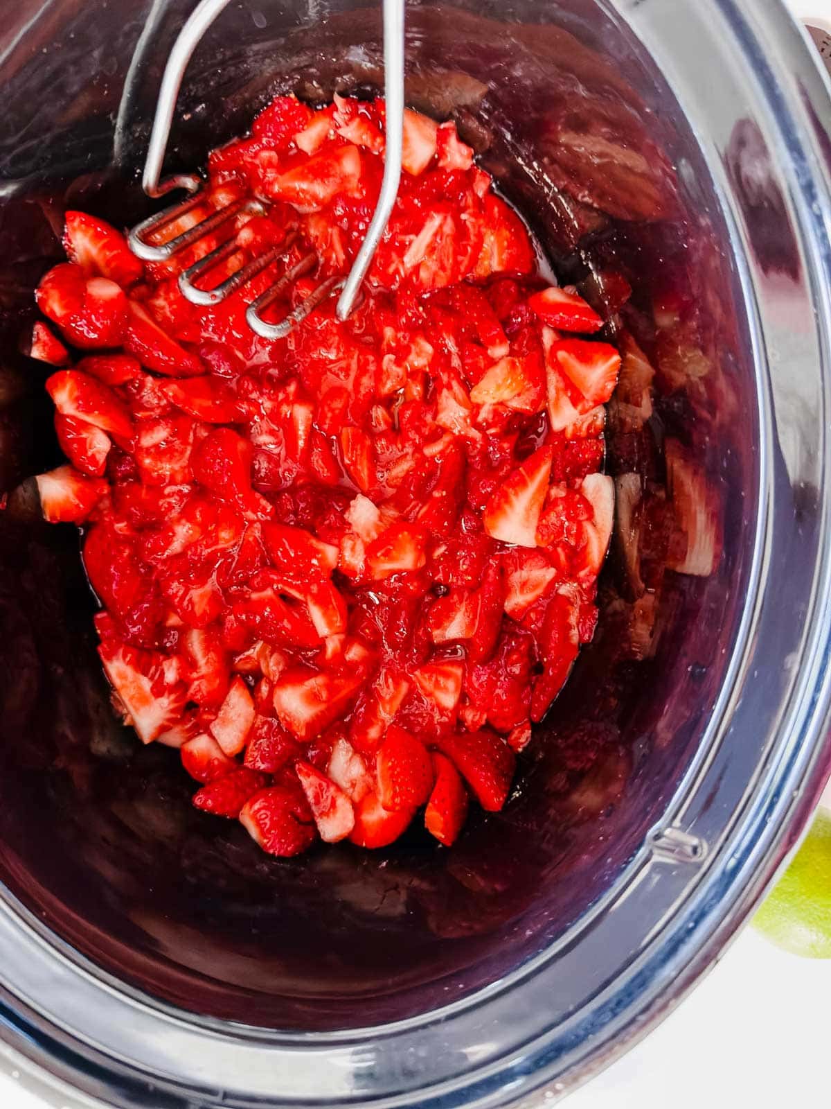 Sliced strawberries being mashed with a metal potato masher inside a black slow cooker or crockpot.
