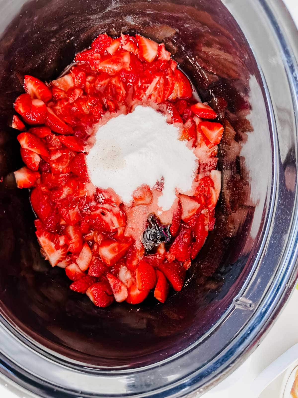 Chopped strawberries with a mound of white sugar and a splash of vanilla extract in a slow cooker, ready to be mixed.