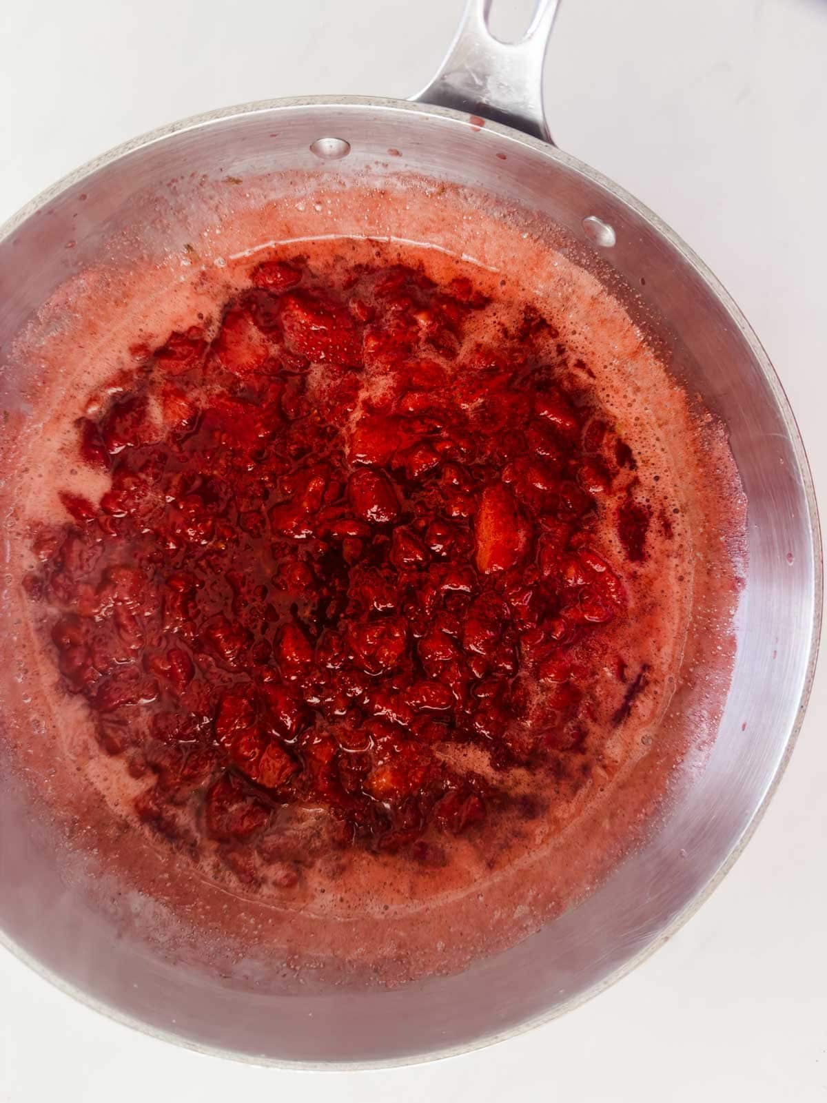 A metal saucepan filled with bubbling, bright red strawberry sauce or jam, with chunks of fruit visible, sits on a white surface.