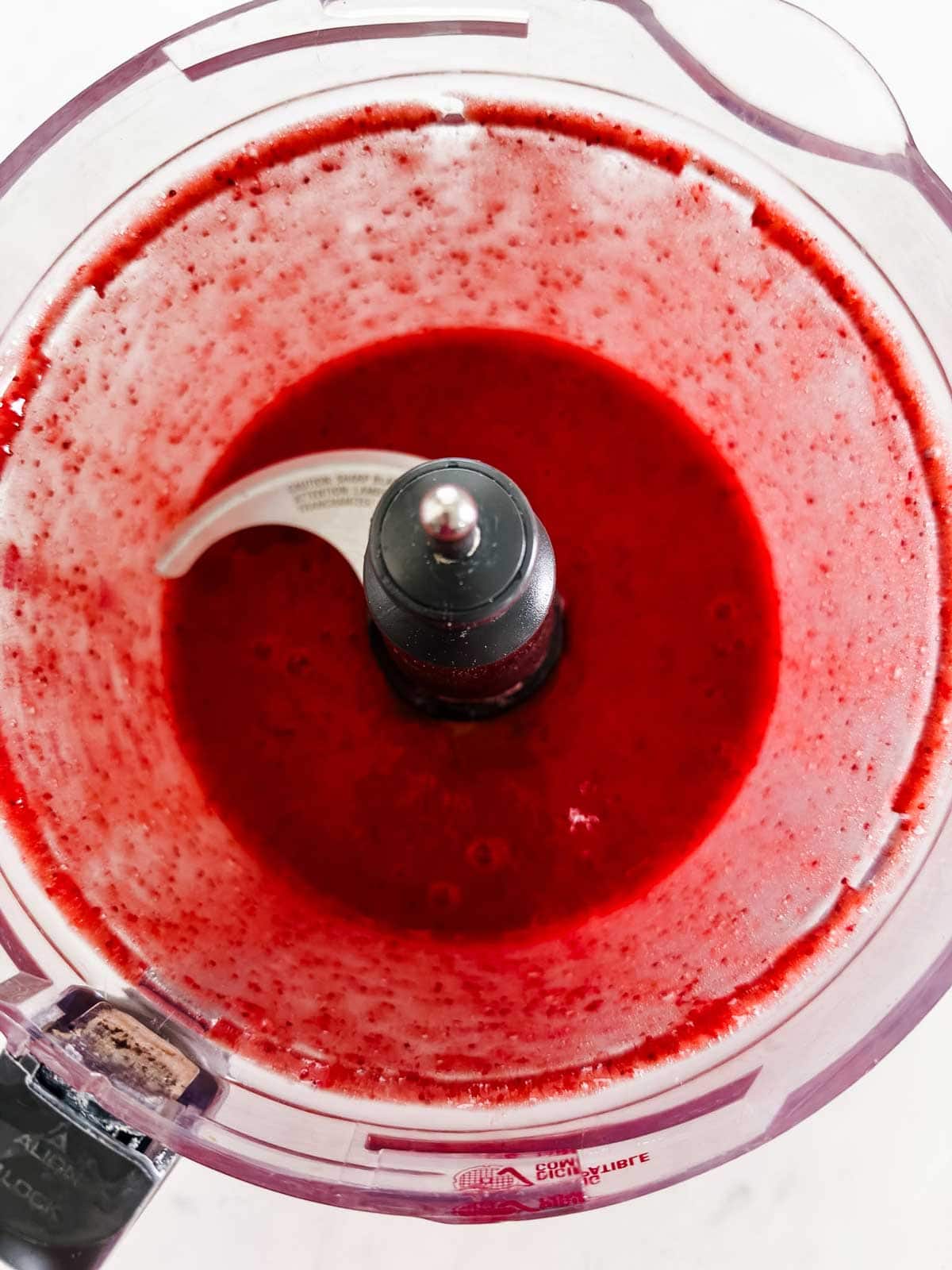 A food processor filled with bright red, blended liquid, likely a fruit puree, viewed from above. The blade is visible in the center of the transparent container.