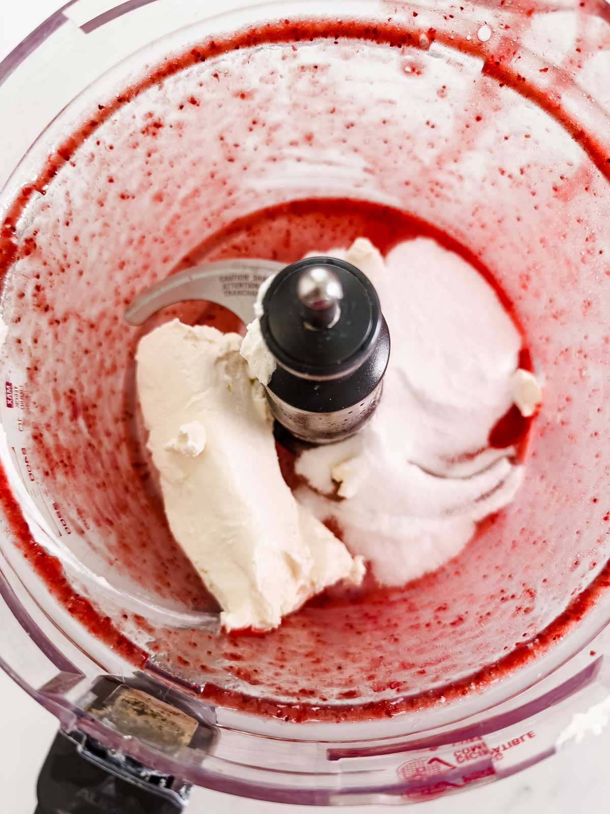 A food processor bowl containing a blend of red berry puree, a block of cream cheese, and a mound of white sugar, ready to be mixed together.