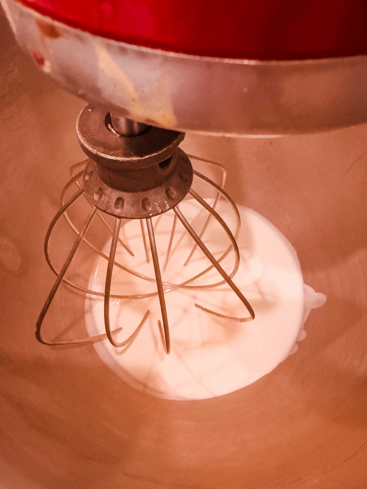 A close-up of a stand mixer with a wire whisk attachment mixing a small amount of liquid, likely cream, in a metal bowl.