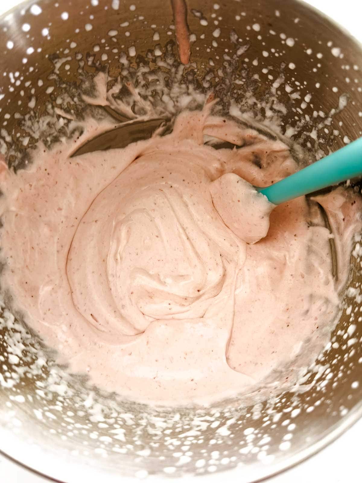 A metal mixing bowl filled with light pink, creamy batter being stirred with a teal spatula. Tiny flecks are visible throughout the mixture, and some batter is splattered on the sides of the bowl.