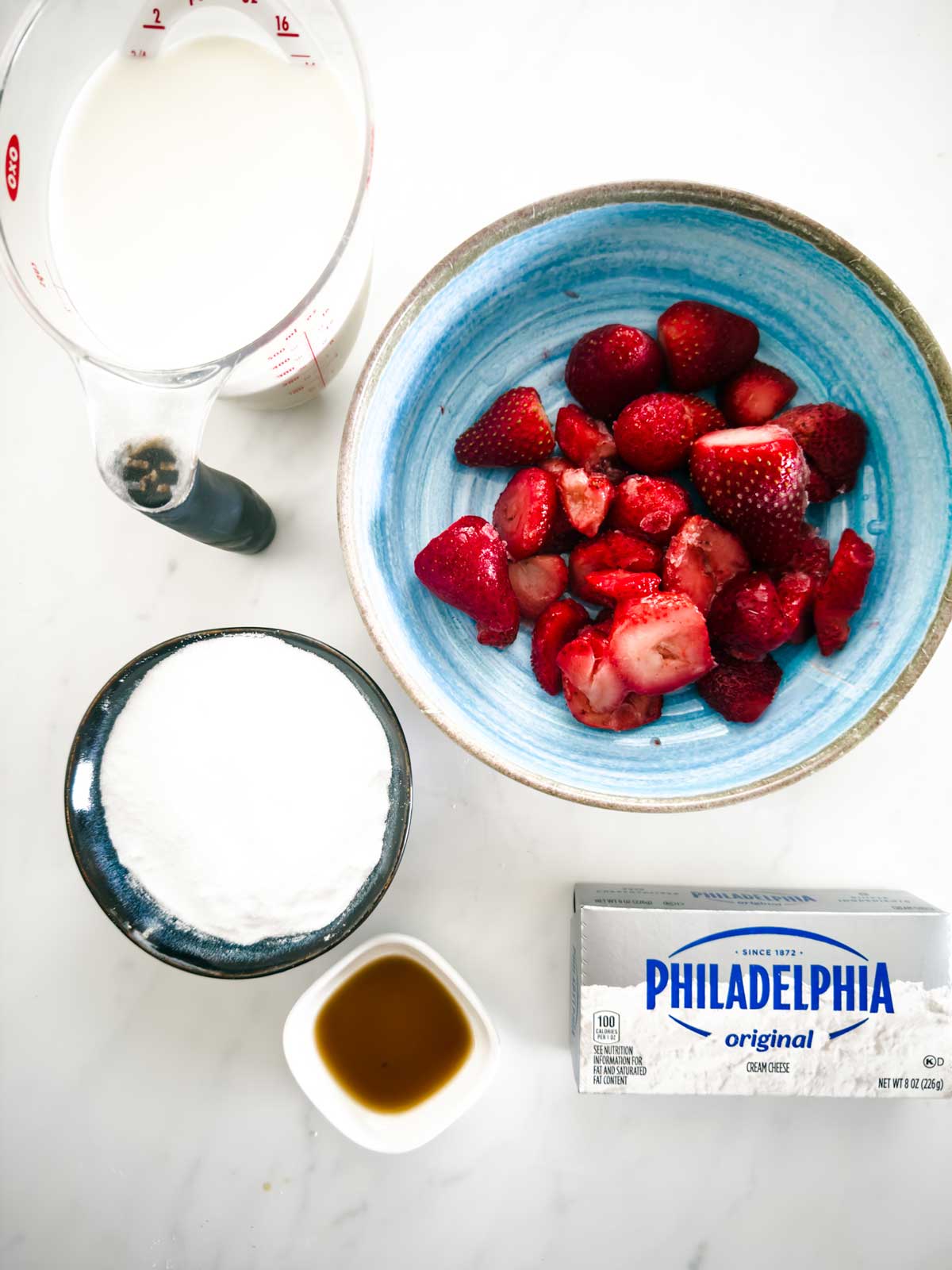 Top-down view of ingredients on a white surface: a blue bowl of halved strawberries, a measuring cup of milk, a cup of sugar, a small dish of vanilla extract, and a block of Philadelphia cream cheese.