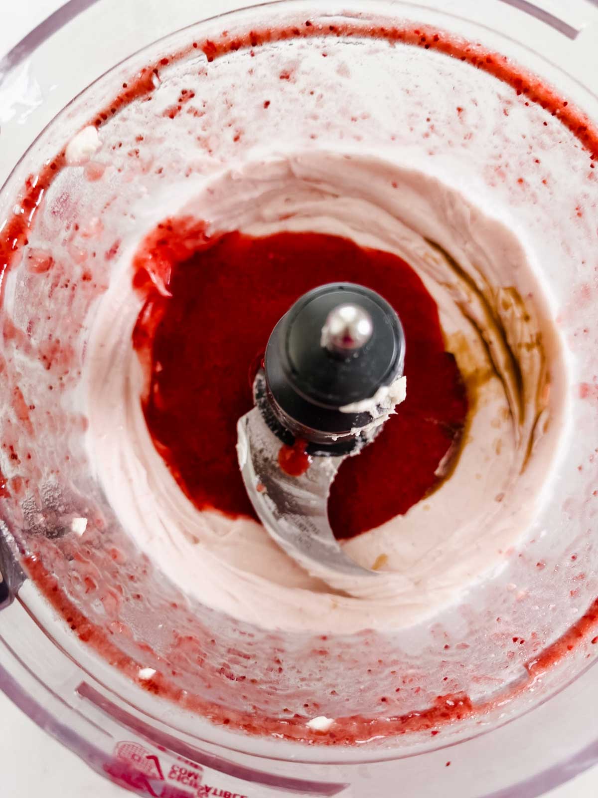 A close-up view of a food processor bowl containing a mixture of red and white ingredients with a metal blade visible in the center.